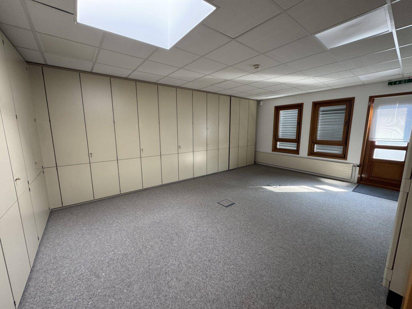 Helles Büro mit Teppichboden, Einbauschränken und zwei Fenstern, mit direktem Zugang nach draußen.
