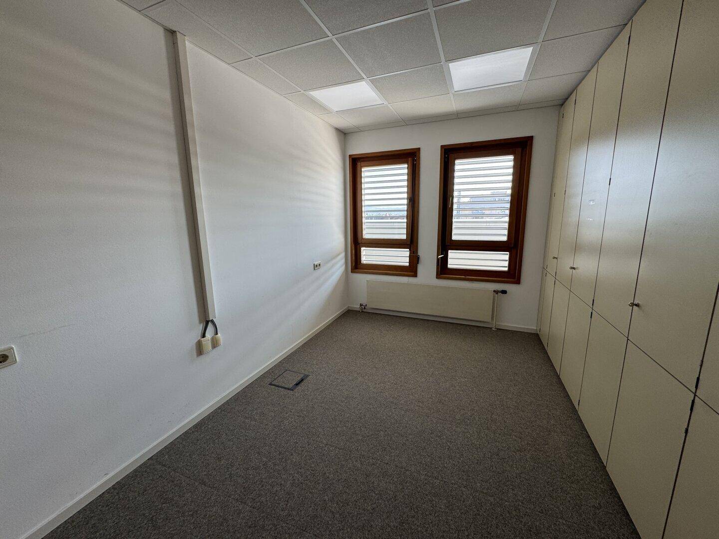 Leeres Büro mit Teppichboden, zwei Fenstern und integrierten Schränken, bietet eine funktionale Arbeitsumgebung.