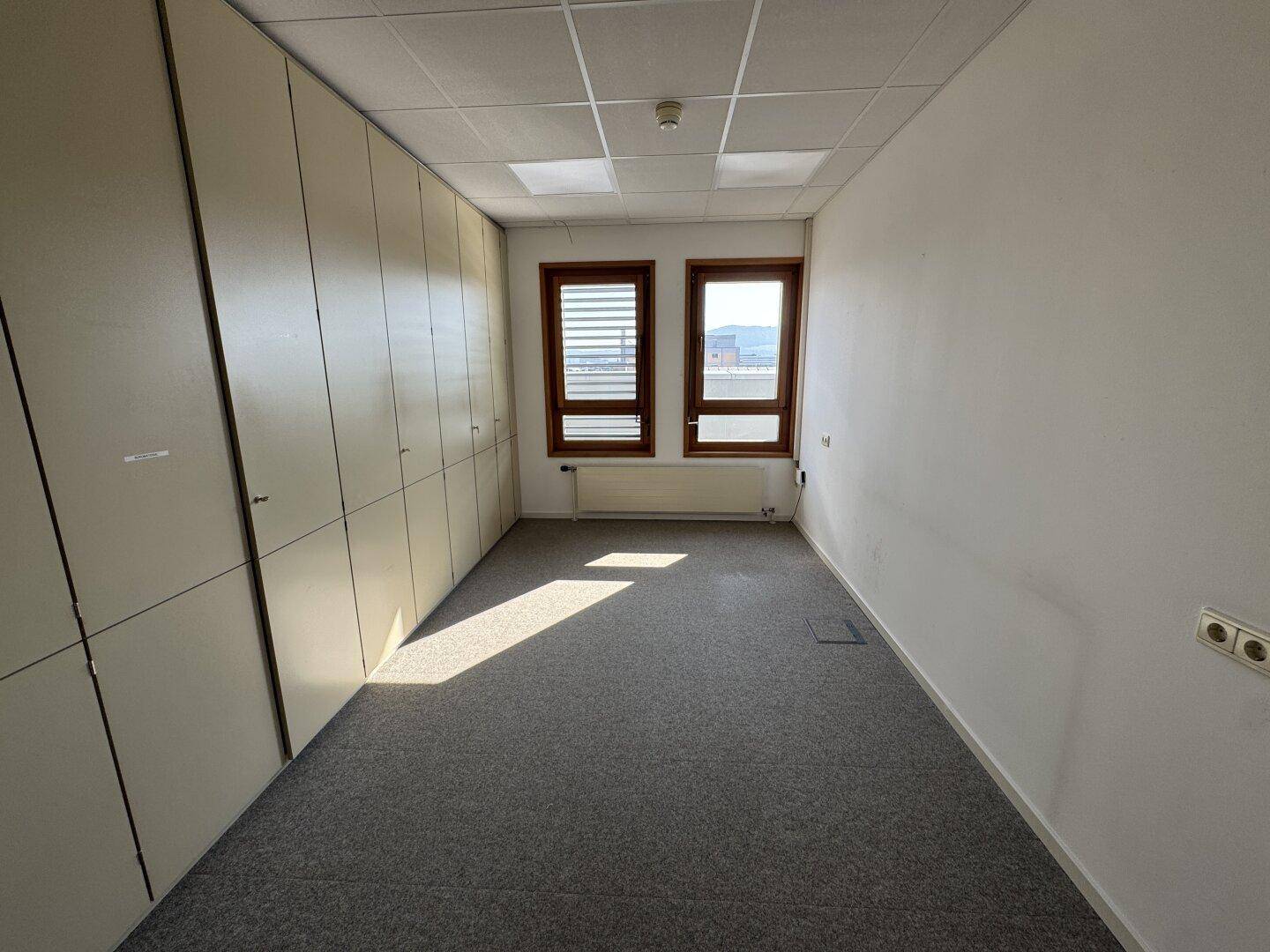 Helles Büro mit Teppichboden, zwei Fenstern und Einbauschränken, bietet einen weiten Blick und viel Tageslicht.