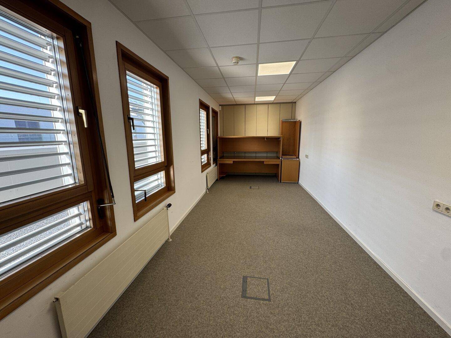 Funktionales Büro mit Teppichboden, Fenstern und einer integrierten Schreibtisch- und Stauraumlösung.