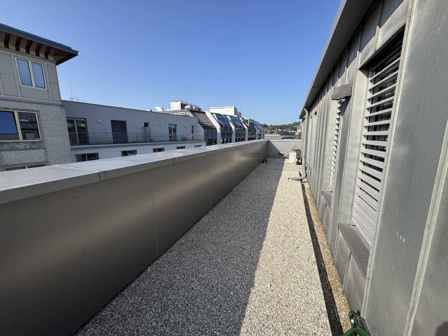 Schmale Dachterrasse mit Kiesboden und niedriger Brüstung, bietet einen Blick auf umliegende Gebäude.