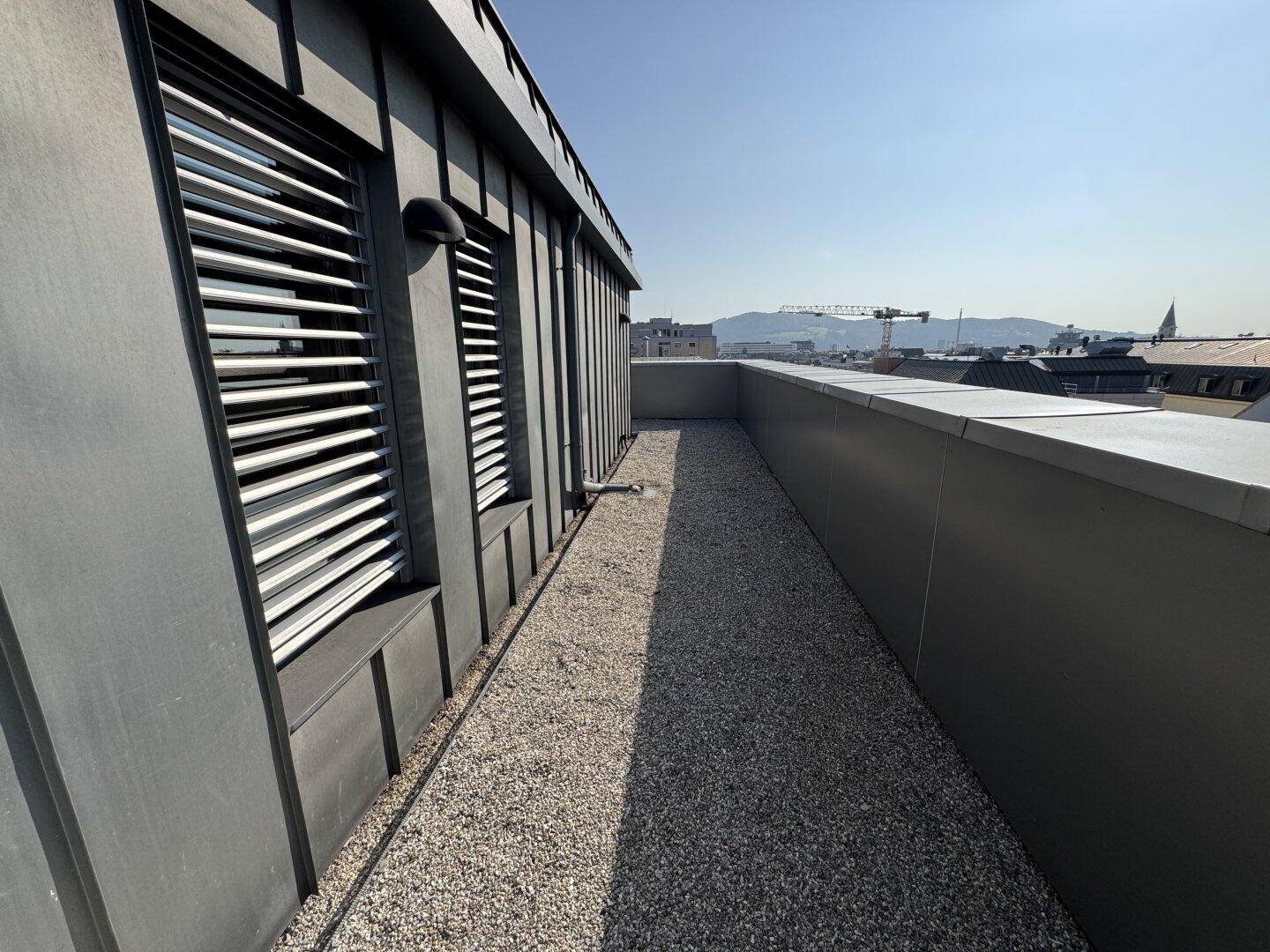 Schmale Dachterrasse mit Kiesboden und Blick auf die Stadt und ferne Berge, ideal für eine kleine Auszeit.