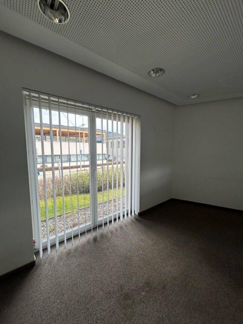 Helles Büro mit dunklem Teppichboden, weißen Wänden, abgehängter Decke und einem großen Fenster mit Jalousien.