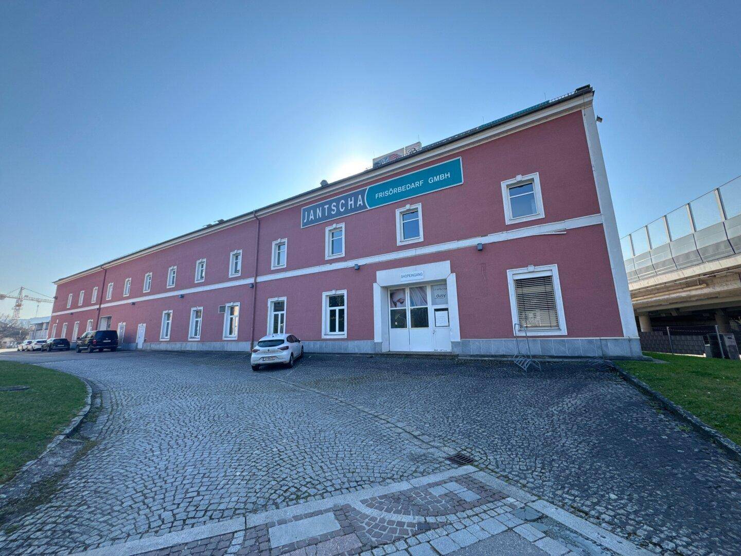 Nahaufnahme der rosafarbenen Gebäudefassade mit weißen Fenstern und einem Eingangsbereich, umgeben von einem gepflasterten Vorplatz.