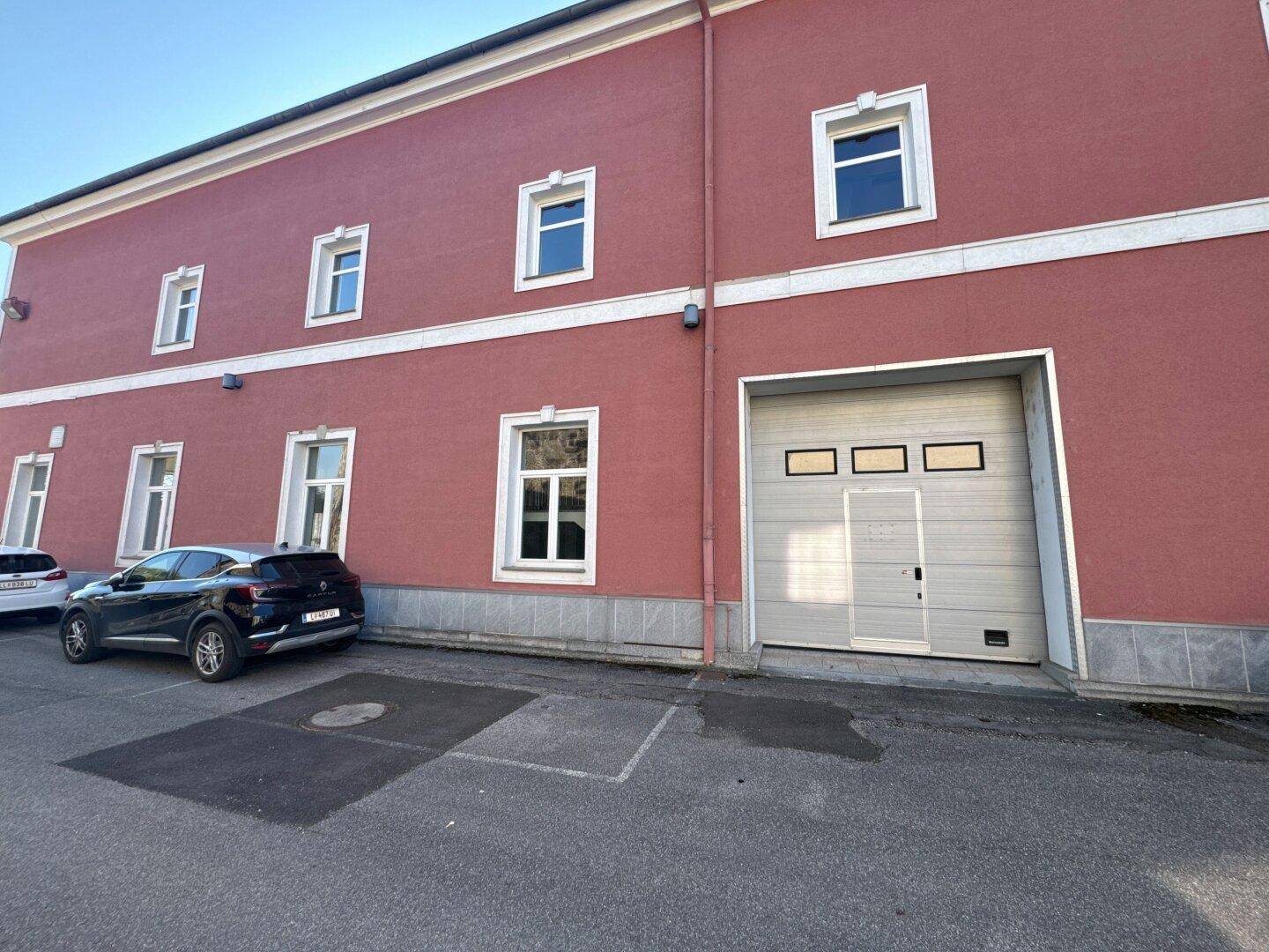 Detailansicht der rosafarbenen Gebäudefassade mit einem großen Rolltor und mehreren Fenstern, davor ein geparktes Auto.
