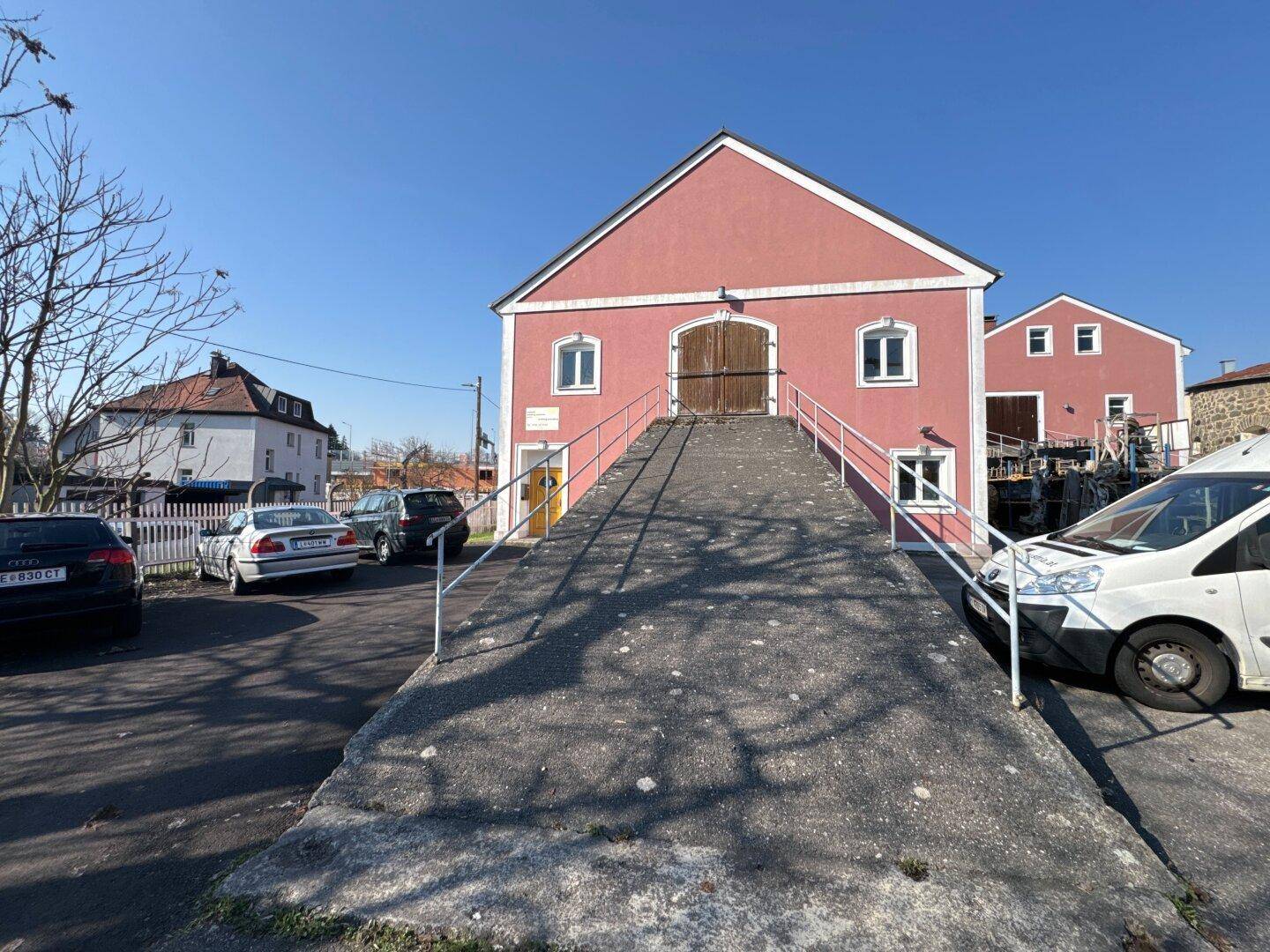 Ansicht des Giebels eines rosafarbenen Gebäudes mit einer langen Zufahrtsrampe und Holztüren, umgeben von Parkplätzen.