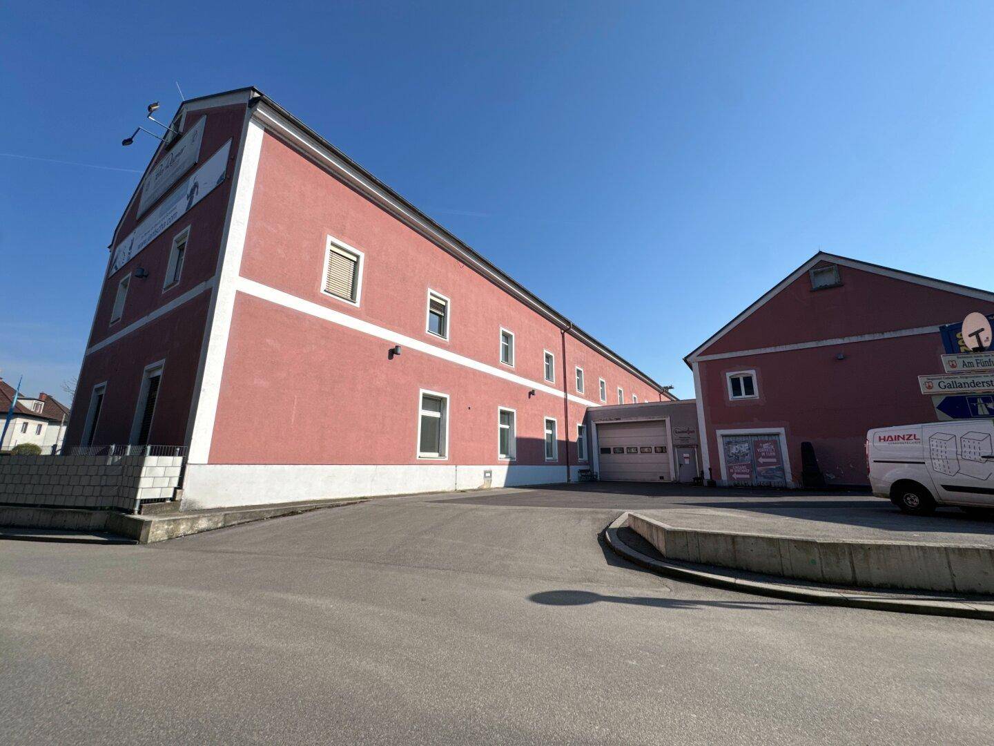 Schräge Ansicht der rosafarbenen Gebäudefassade mit einem Garagentor und einem gepflasterten Zufahrtsbereich unter blauem Himmel.