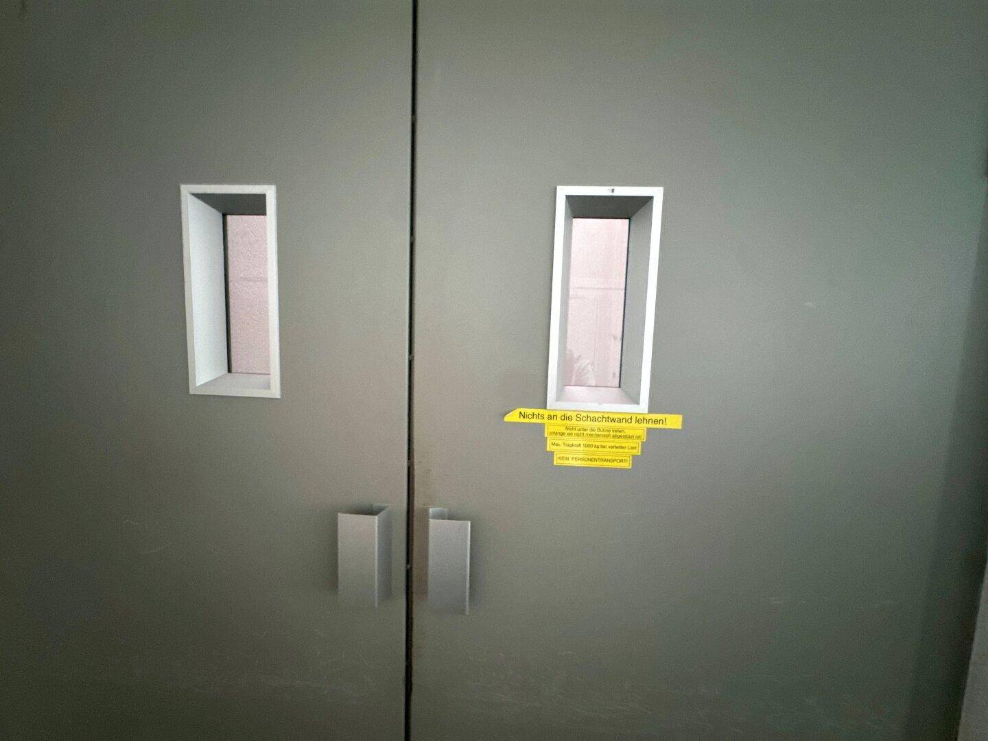 Detailaufnahme von zwei grauen Aufzugtüren mit kleinen Fenstern und einem gelben Warnhinweis.
