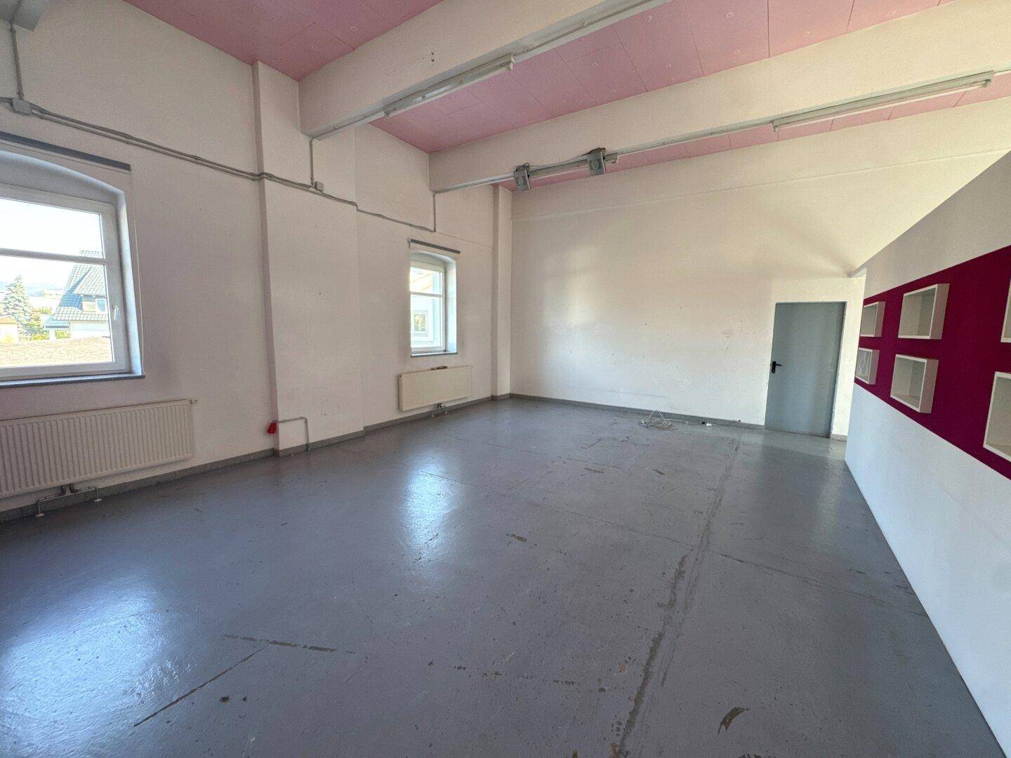 Großer, leerer Raum mit grauen Betonböden, weißen Wänden und rosa Deckenbalken, beleuchtet durch Fenster und Neonröhren.