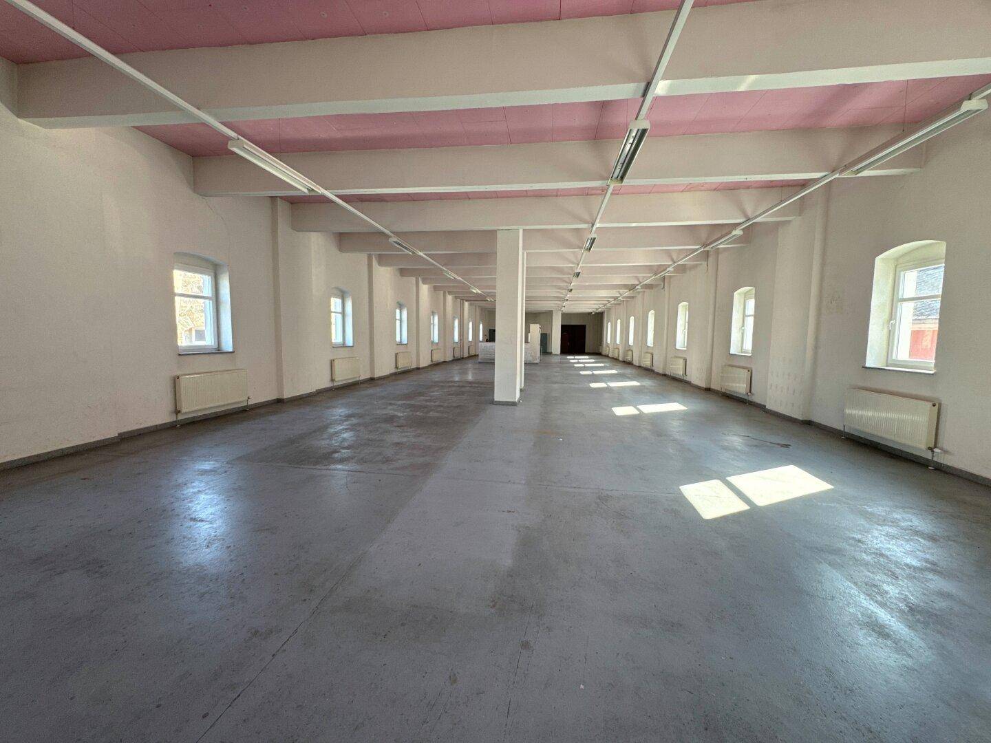 Langer, offener Raum mit grauen Betonböden, weißen Wänden und rosa Deckenbalken, beleuchtet durch zahlreiche Fenster.
