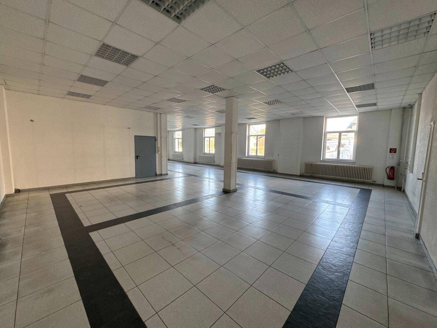 Großer, offener Büroraum mit weißen Fliesenböden und schwarzen Akzenten, abgehängter Decke und Fenstern.