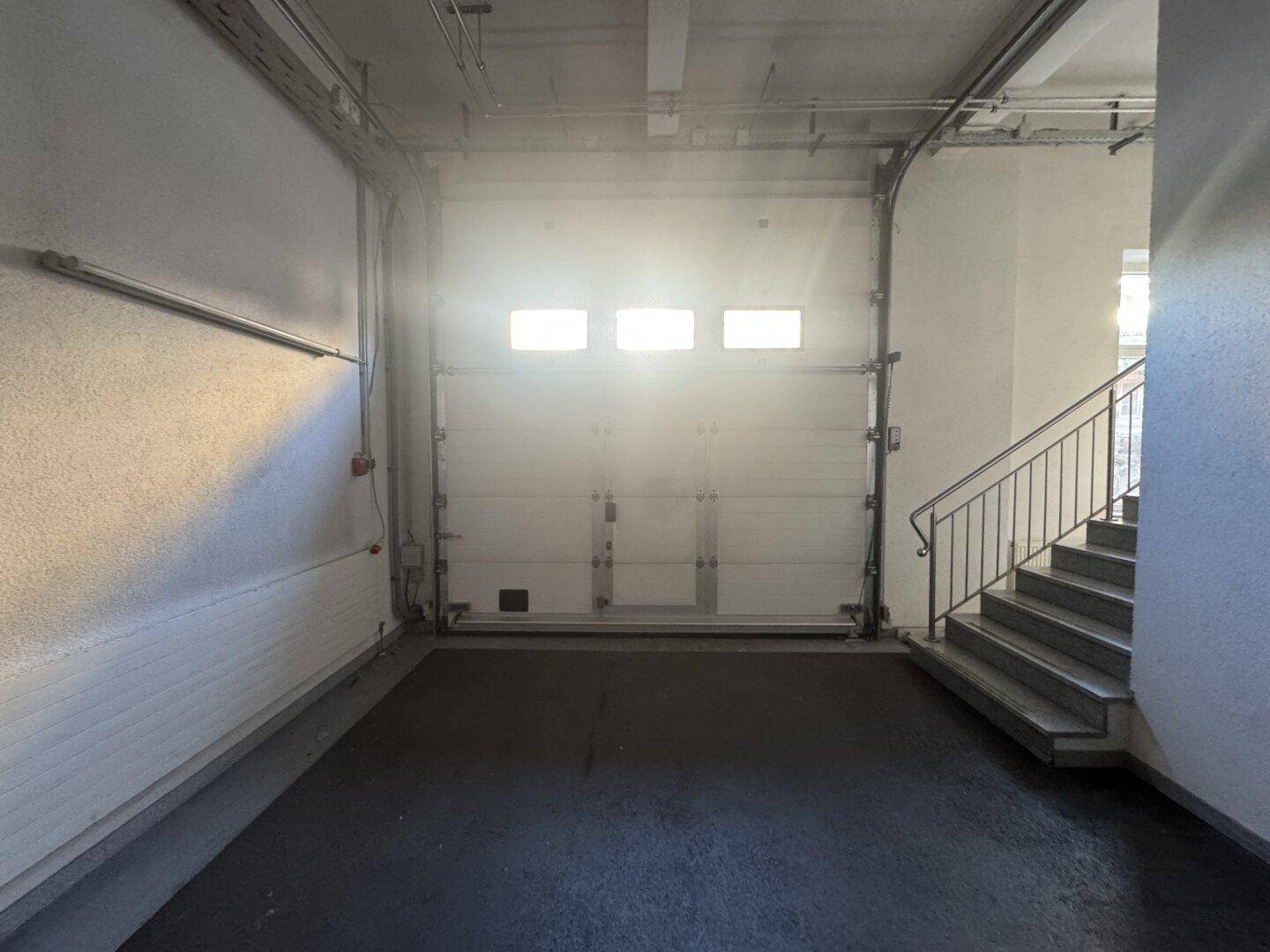 Innenansicht einer Garage mit einem großen Rolltor, dunklem Boden und einer Treppe, die nach oben führt.