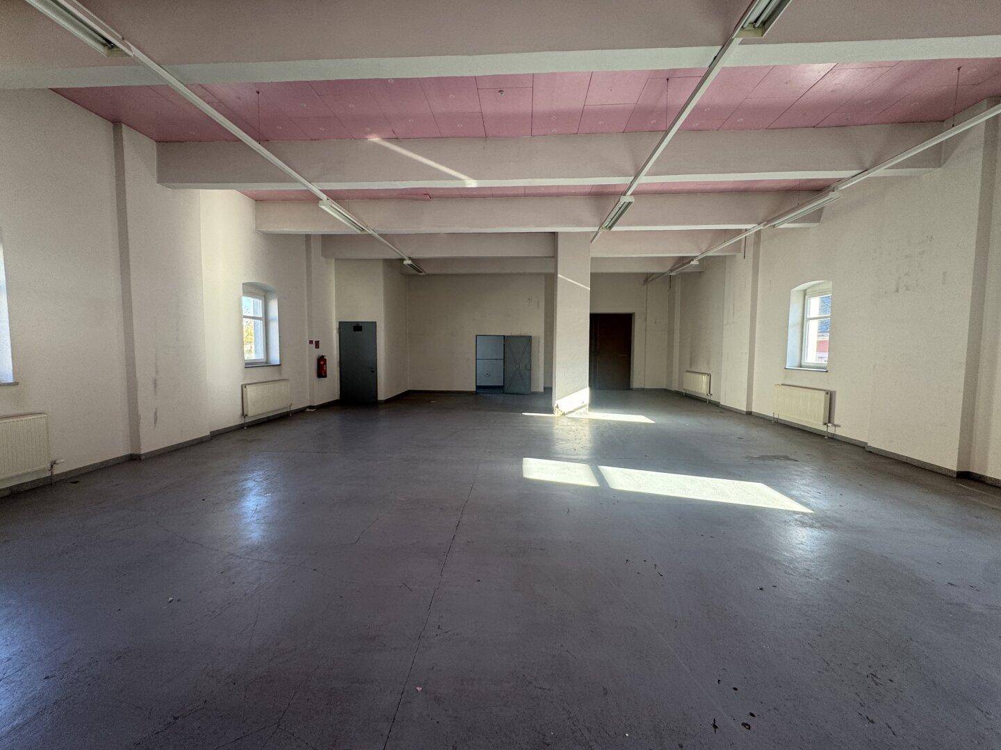Geräumiger, leerer Raum mit grauen Betonböden, weißen Wänden und rosa Deckenbalken, beleuchtet durch Fenster.