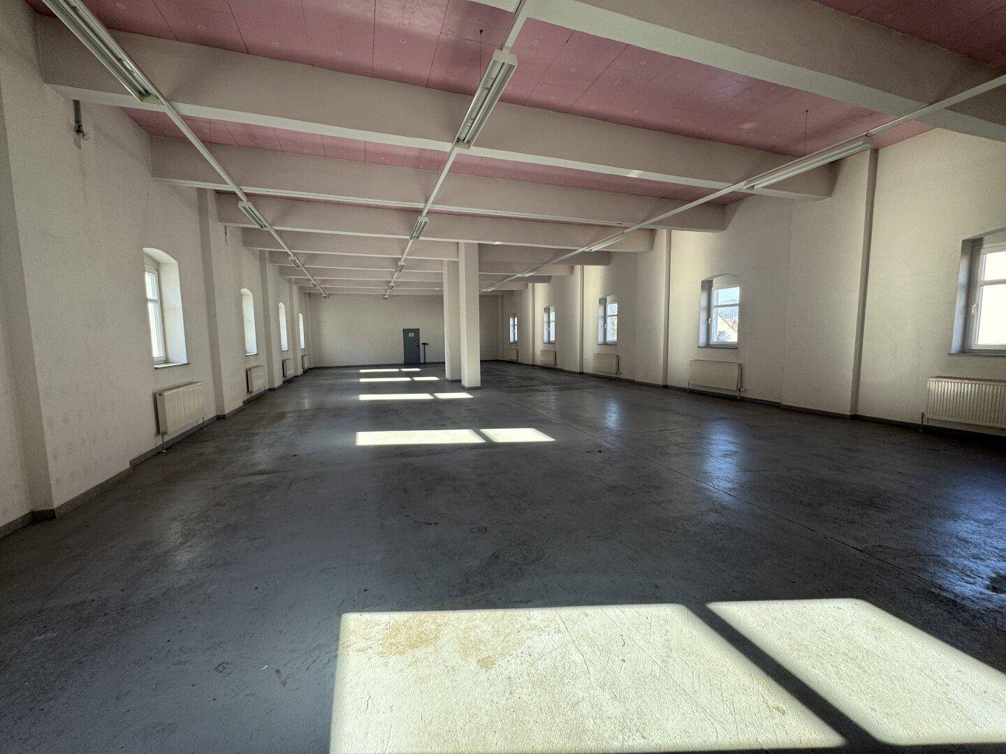 Langer, offener Raum mit grauen Betonböden, weißen Wänden und rosa Deckenbalken, durch Fenster hell erleuchtet.