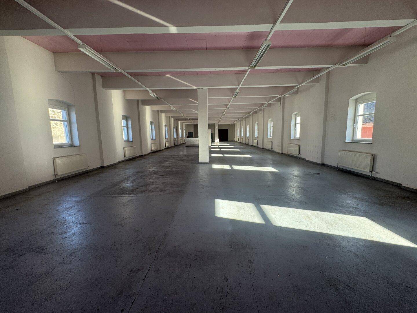Weitläufiger, leerer Raum mit grauen Betonböden, weißen Wänden und rosa Deckenbalken, durch Fenster mit Tageslicht versorgt.