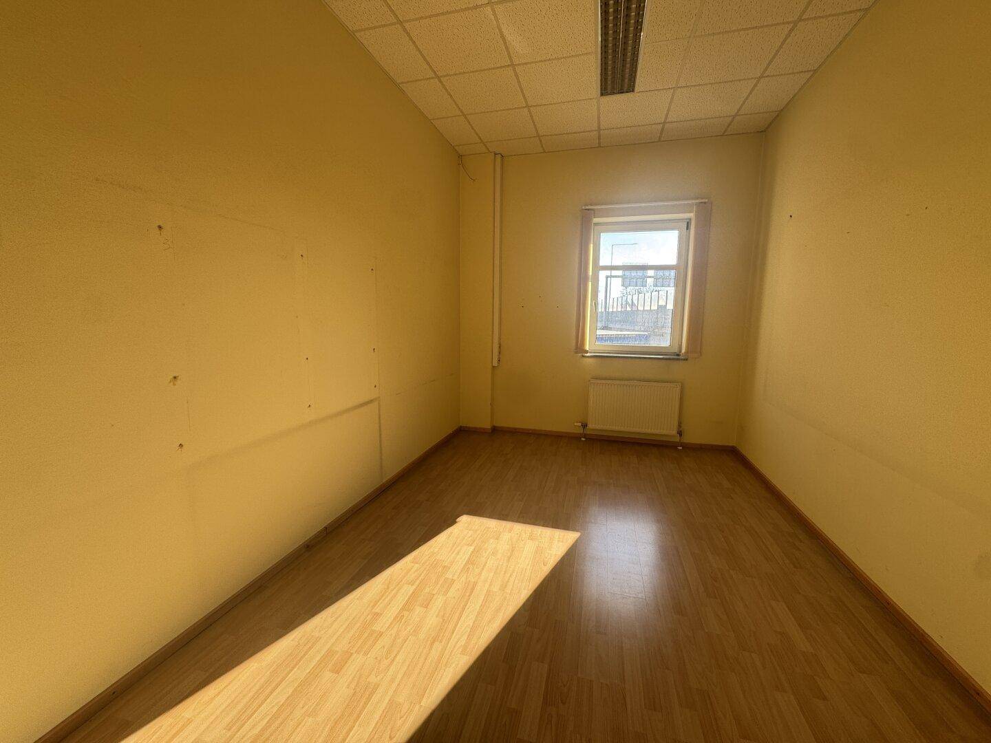 Kleiner, leerer Büroraum mit gelben Wänden, Holzfußboden und einem Fenster, das Tageslicht hereinlässt.