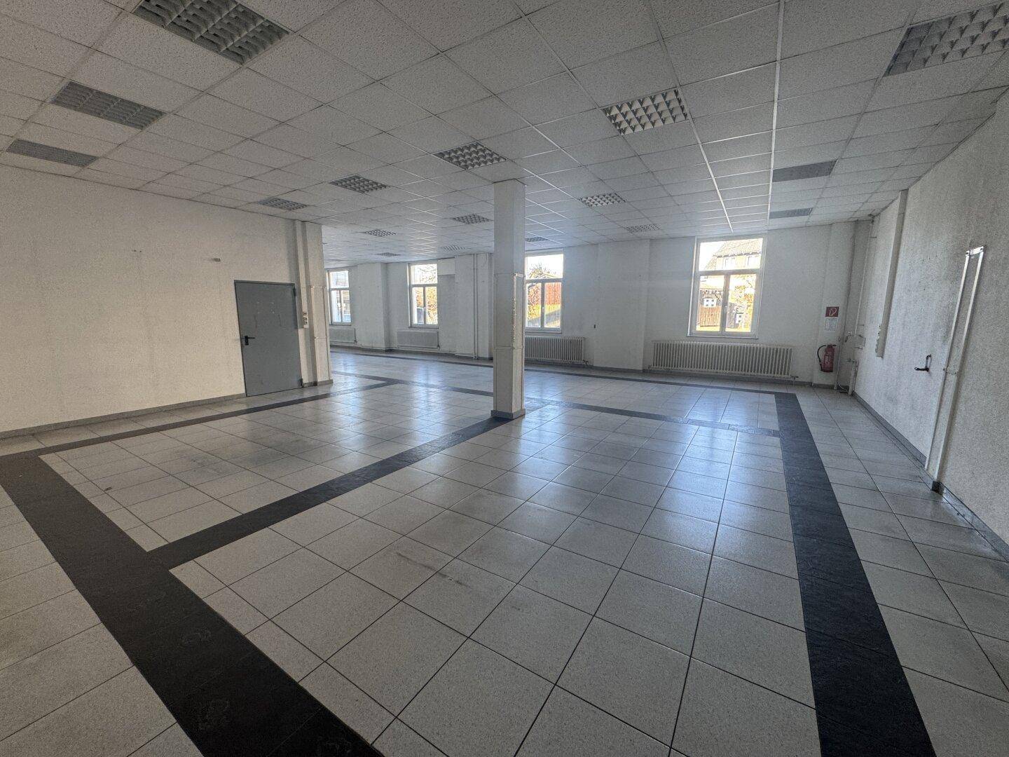 Großer, offener Büroraum mit weißen Fliesenböden und schwarzen Akzenten, abgehängter Decke und Fenstern.