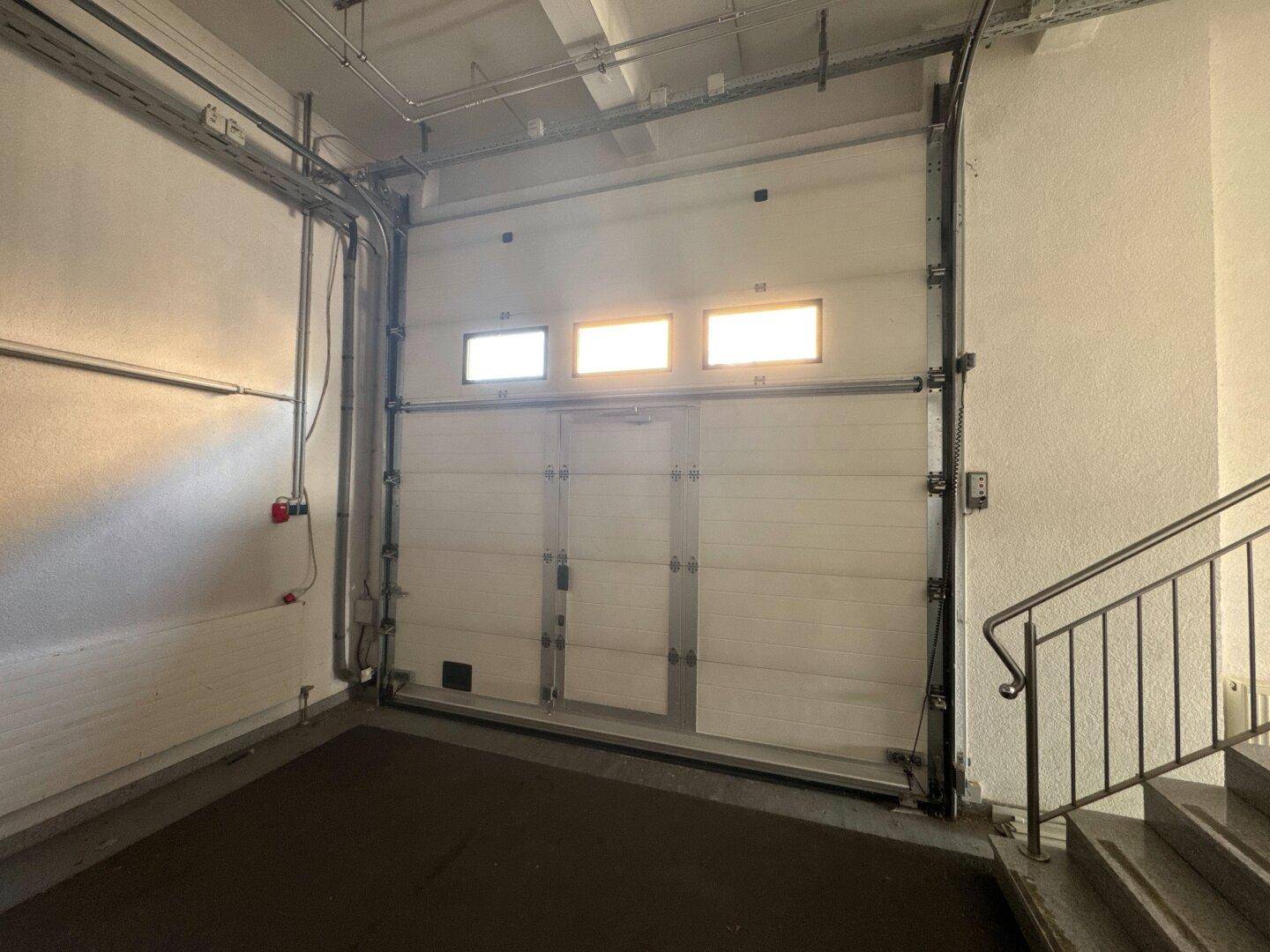 Innenansicht einer Garage mit einem großen Rolltor, dunklem Boden und einer Treppe, die nach oben führt.
