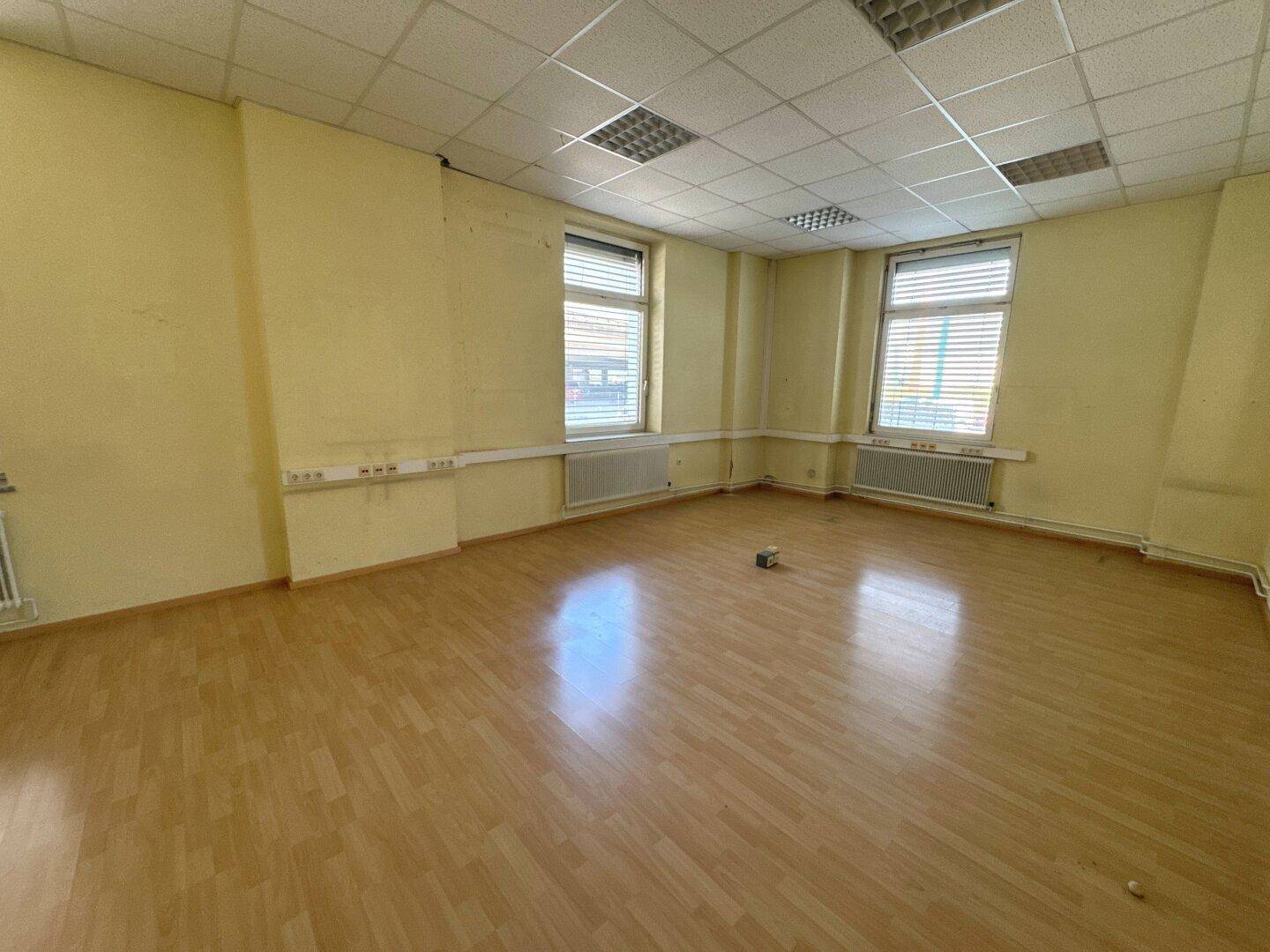 Geräumiger Büroraum mit gelben Wänden, Holzfußboden und zwei Fenstern mit Jalousien, die Tageslicht hereinlassen.