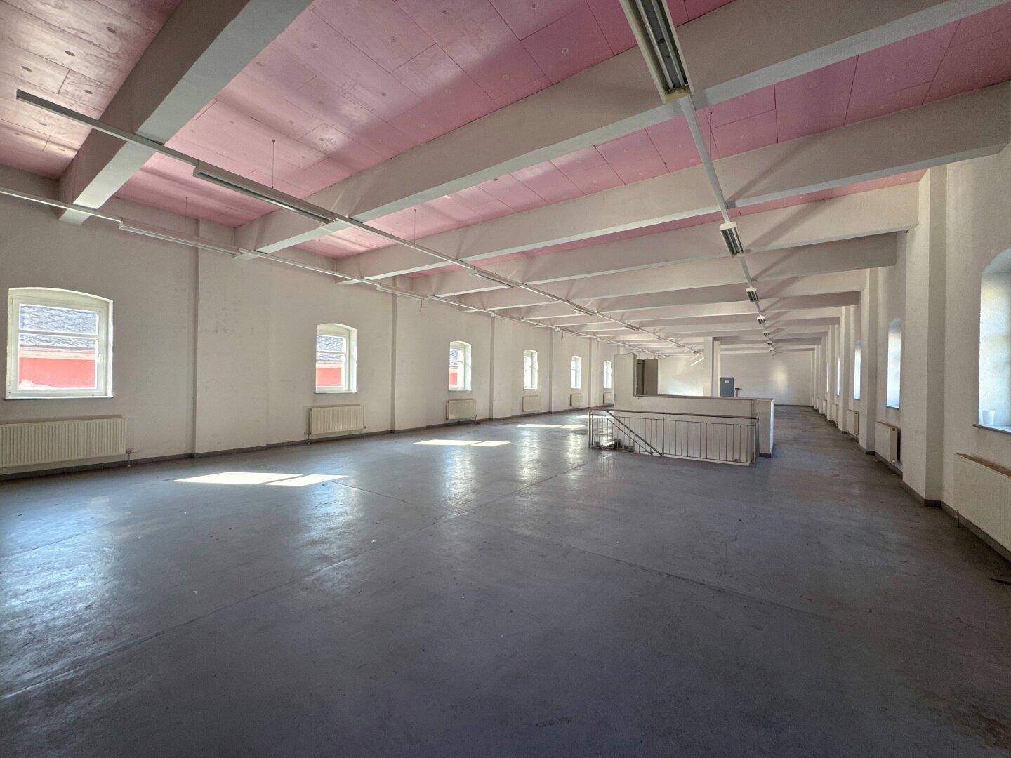 Langer, offener Raum mit grauen Betonböden, weißen Wänden und rosa Deckenbalken, durch Fenster hell erleuchtet.