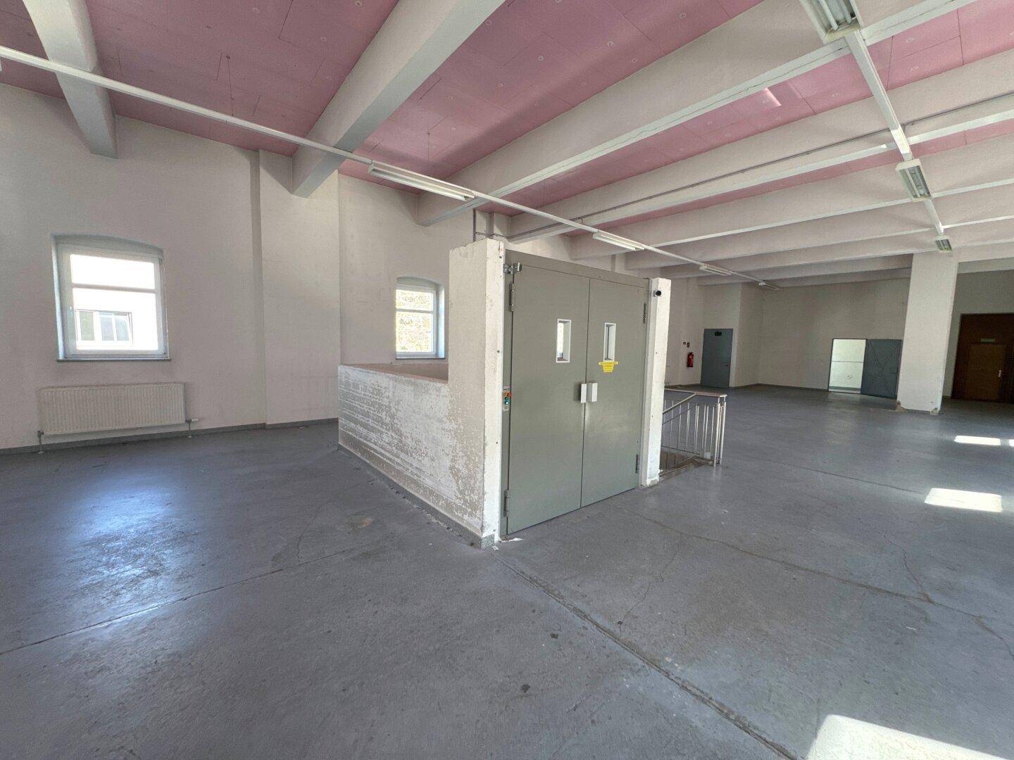 Geräumiger, leerer Raum mit grauen Betonböden, weißen Wänden und rosa Deckenbalken, mit einem Aufzug in der Mitte.
