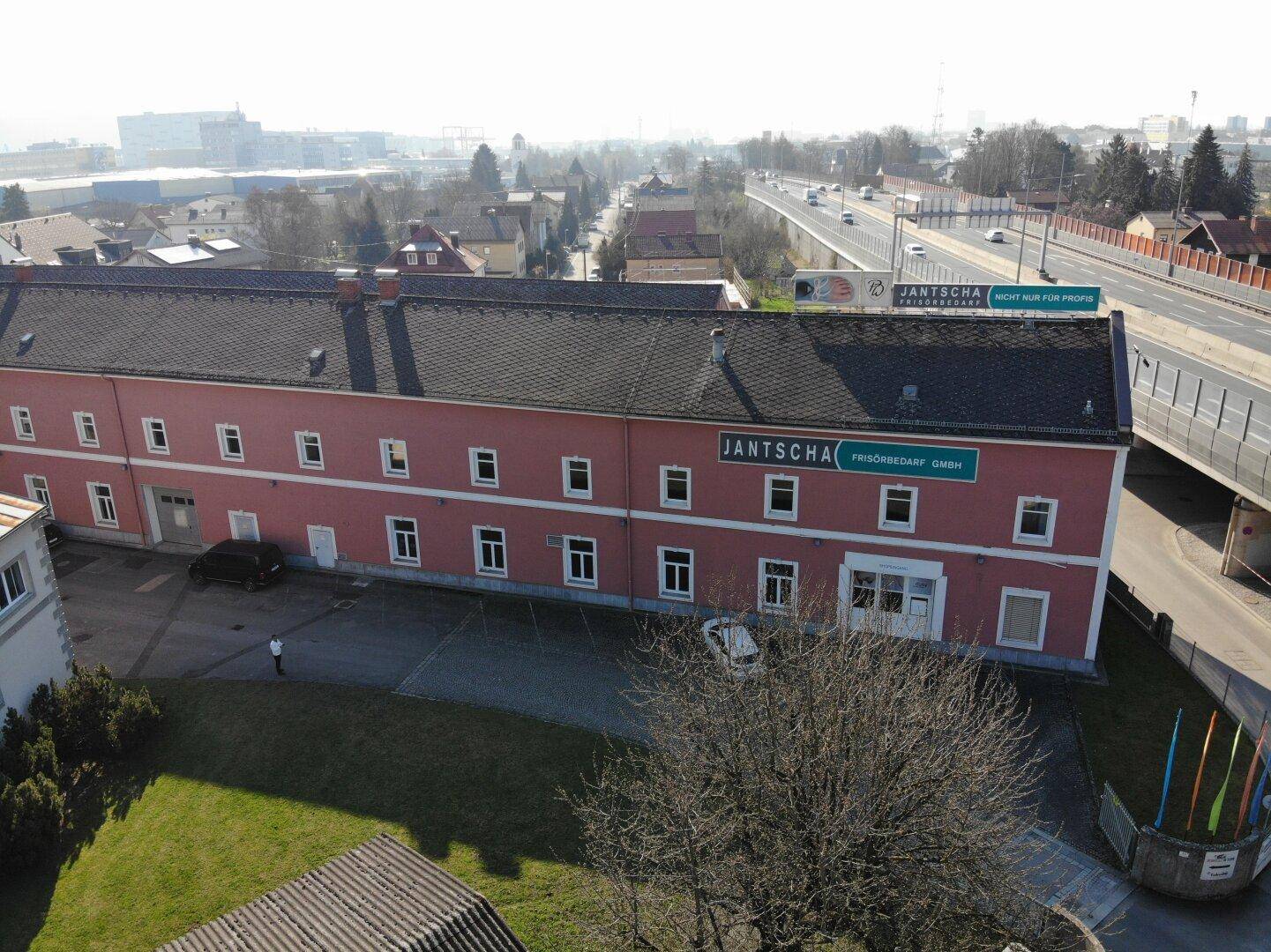 Ansicht der langen rosafarbenen Gebäudefassade mit vielen Fenstern und einem großen Tor, umgeben von Parkplätzen und einer Straße.