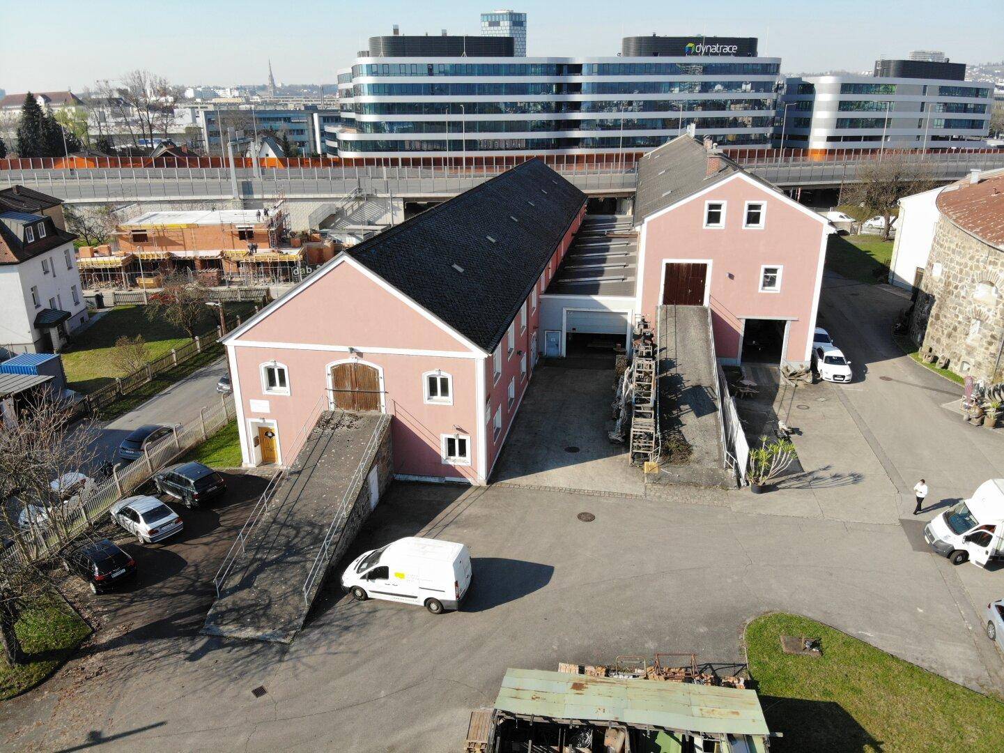 Luftaufnahme der beiden rosafarbenen Gebäude mit Rampen und Parkplätzen, im Hintergrund moderne Bürogebäude und eine Autobahn.