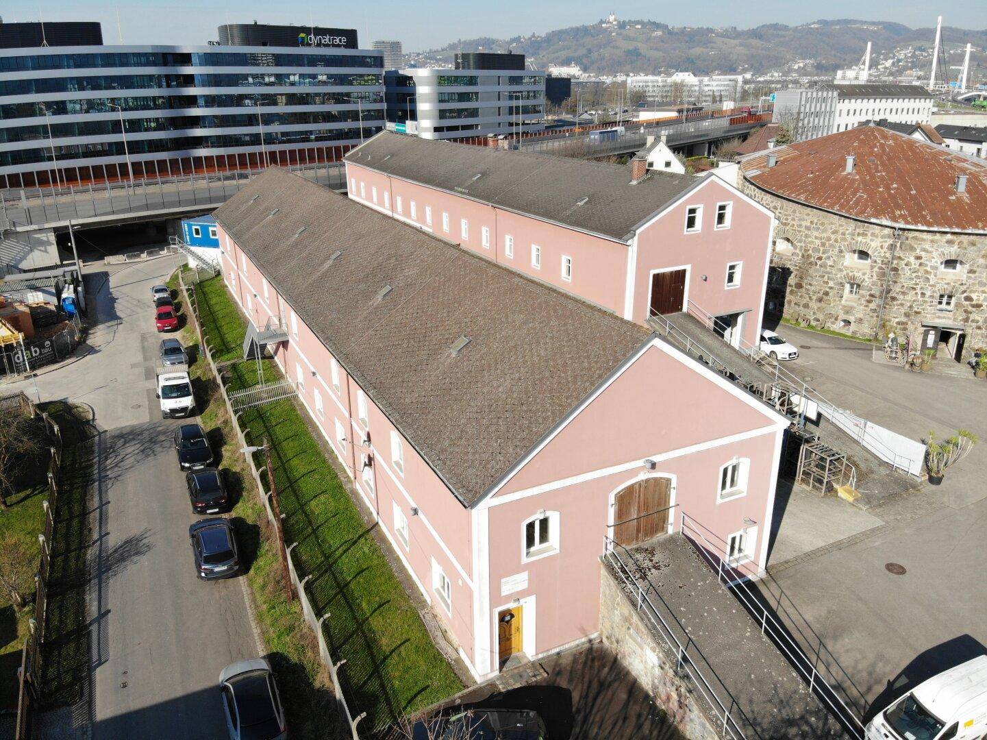 Vogelperspektive auf zwei langgestreckte Gebäude mit rosafarbener Fassade und braunen Dächern, umgeben von Parkplätzen und einer städtischen Umgebung.