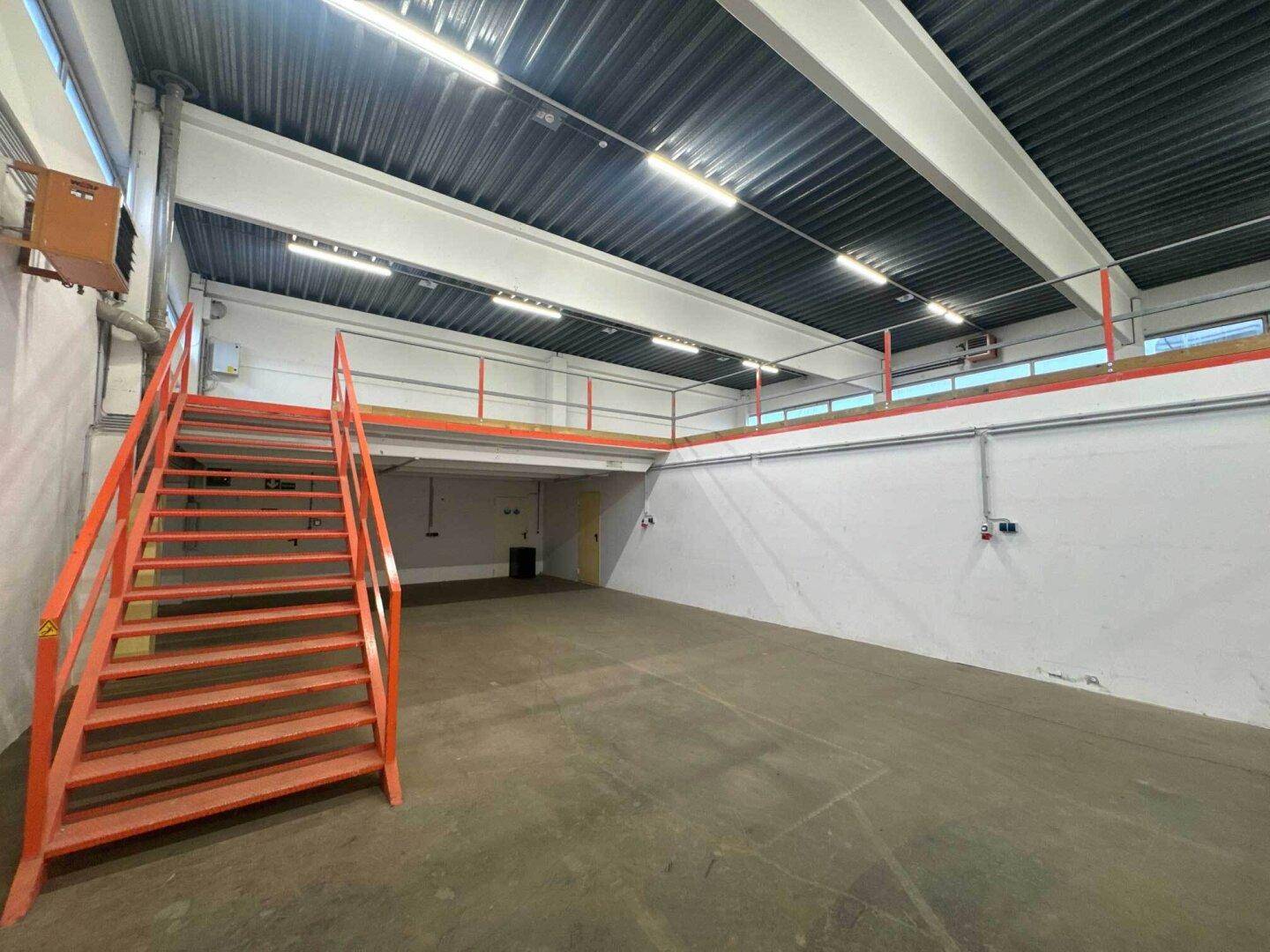 Geräumige Lagerhalle mit hoher Decke, Betonboden und einer orangefarbenen Treppe, die zu einer Empore führt.
