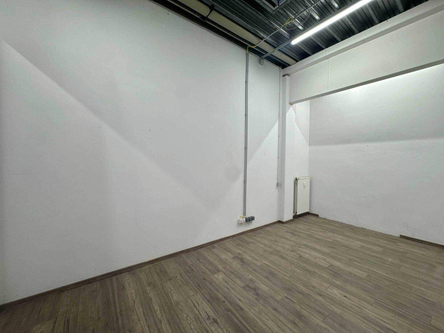 Kleiner, heller Raum mit Holzboden, weißen Wänden und sichtbaren Leitungen unter der Decke.