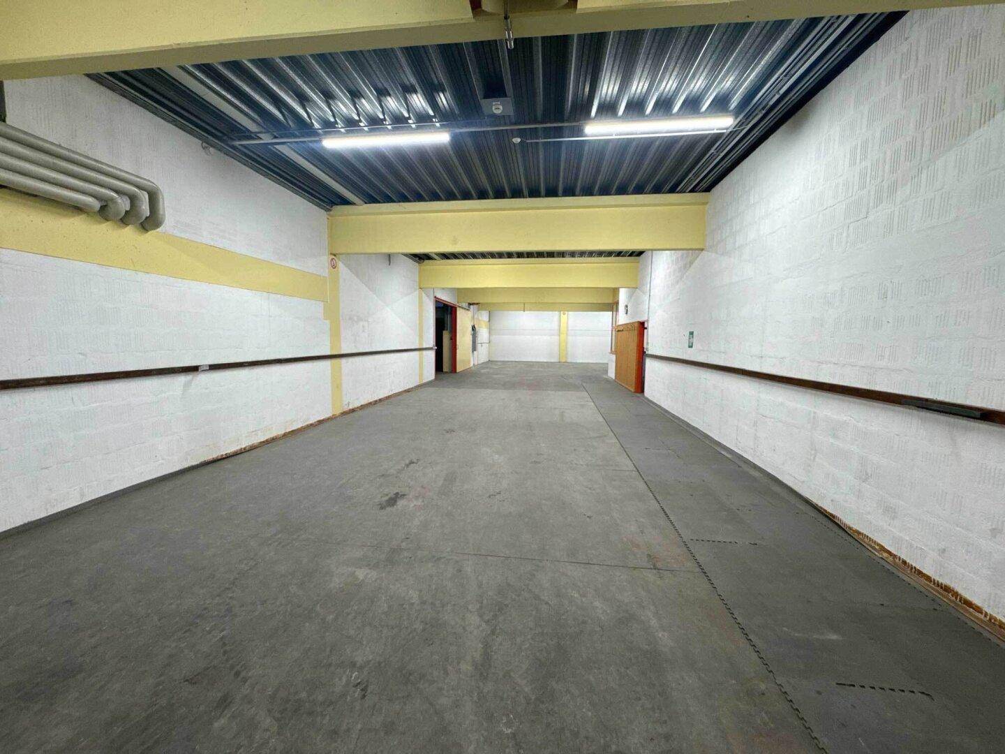 Langer, leerer Flur mit Betonboden, weißen Wänden und gelben Trägern unter der hohen Decke.