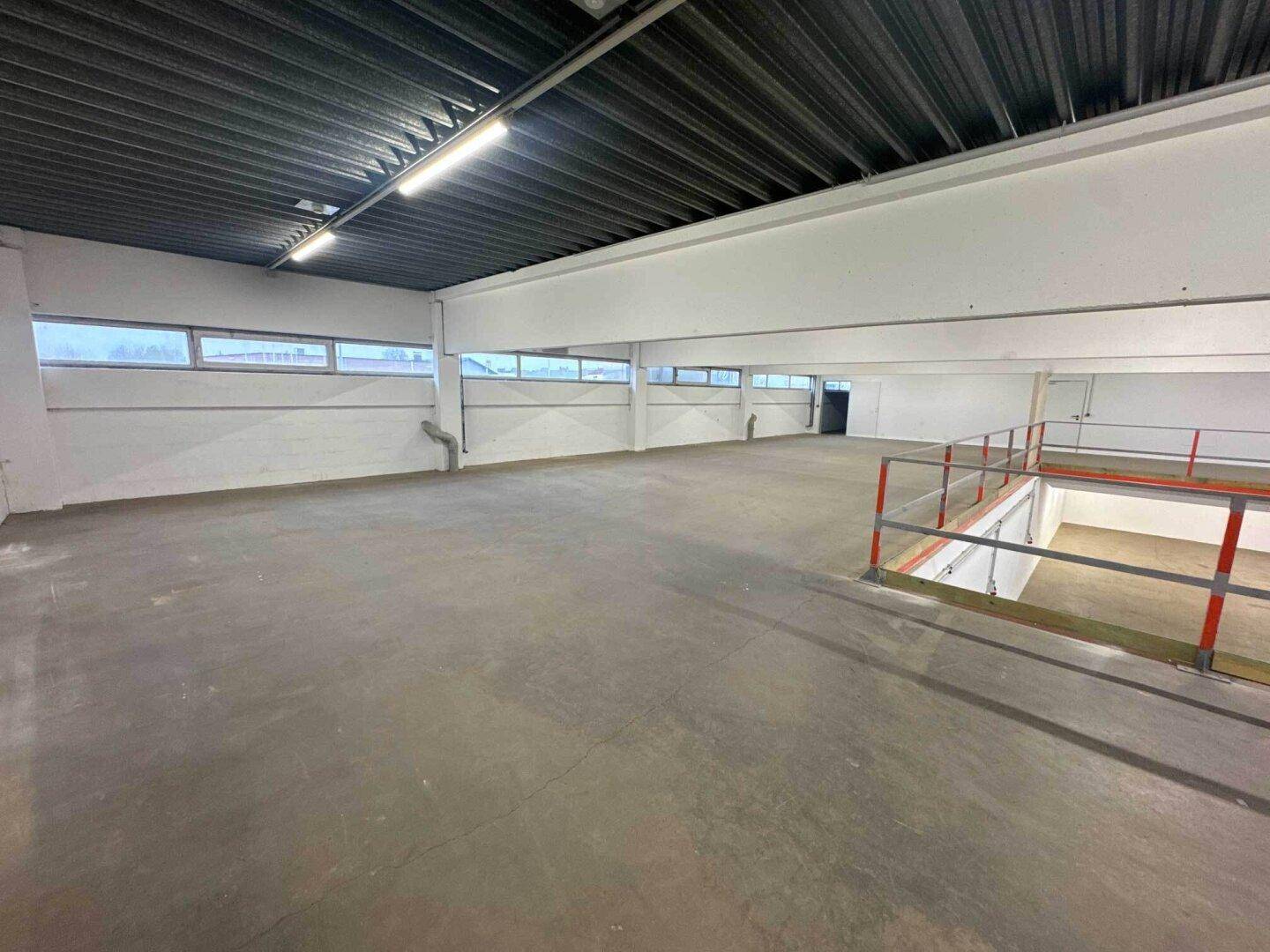 Obere Ebene einer Lagerhalle mit Betonboden, niedrigen Fenstern und Blick auf den unteren Bereich.