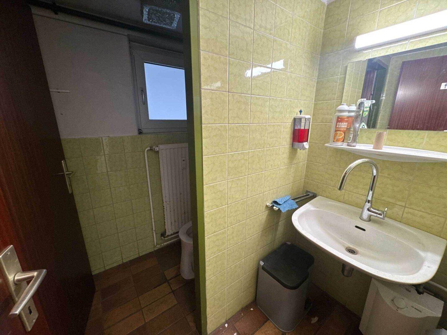 Funktionales Badezimmer mit grünen Fliesen, Waschbecken, Toilette und einem kleinen Fenster.