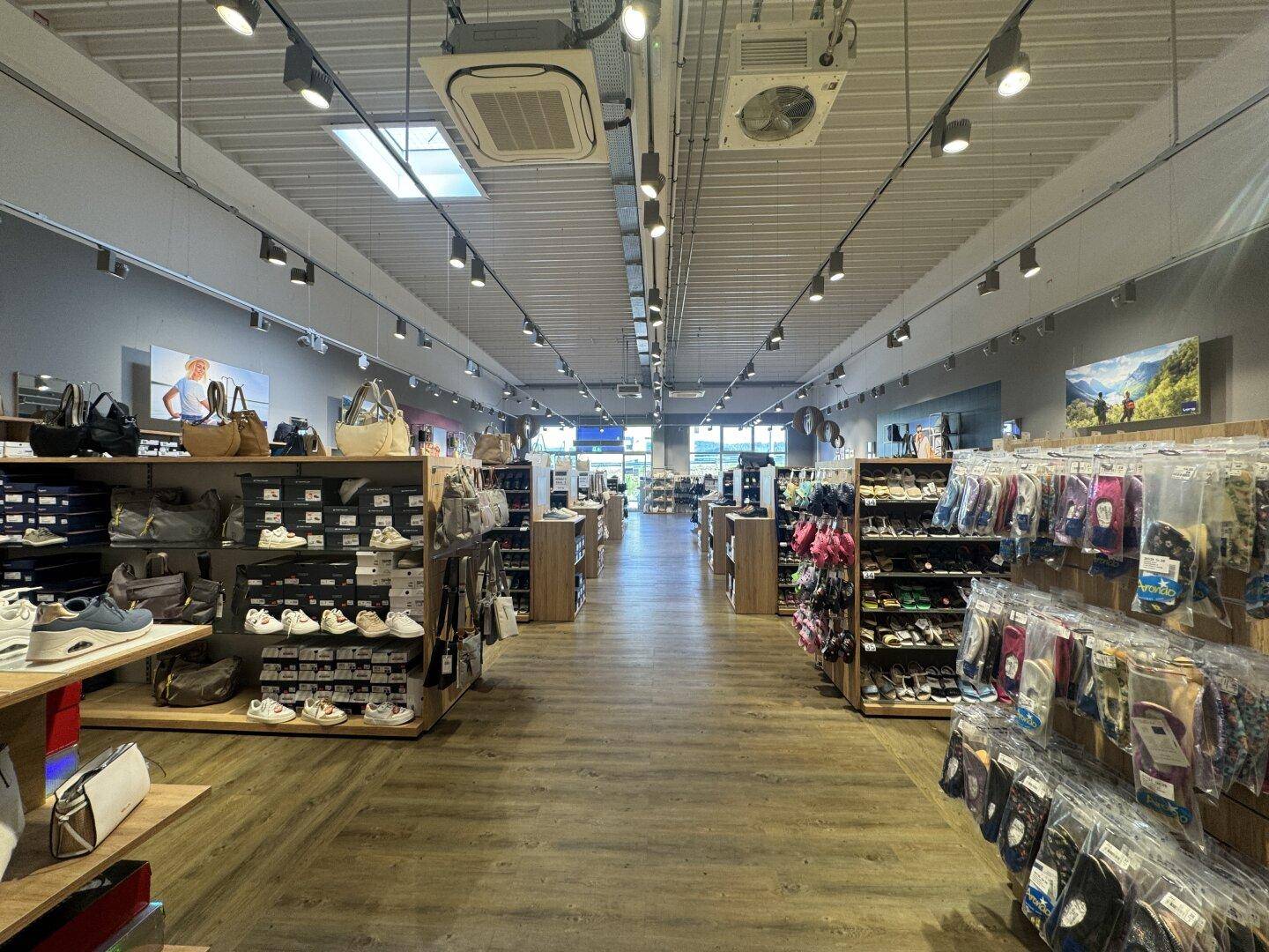Langer Gang in einem Schuhgeschäft mit umfangreicher Warenauslage und Deckenbeleuchtung, die den Raum erhellt.