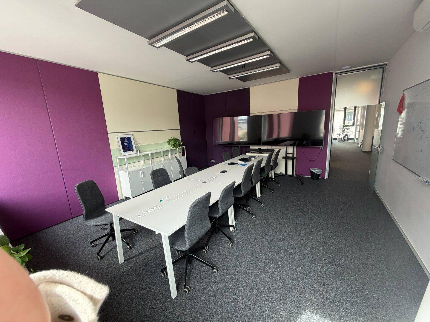 Moderner Konferenzraum mit langem Tisch, ergonomischen Stühlen und großen Bildschirmen für Präsentationen.
