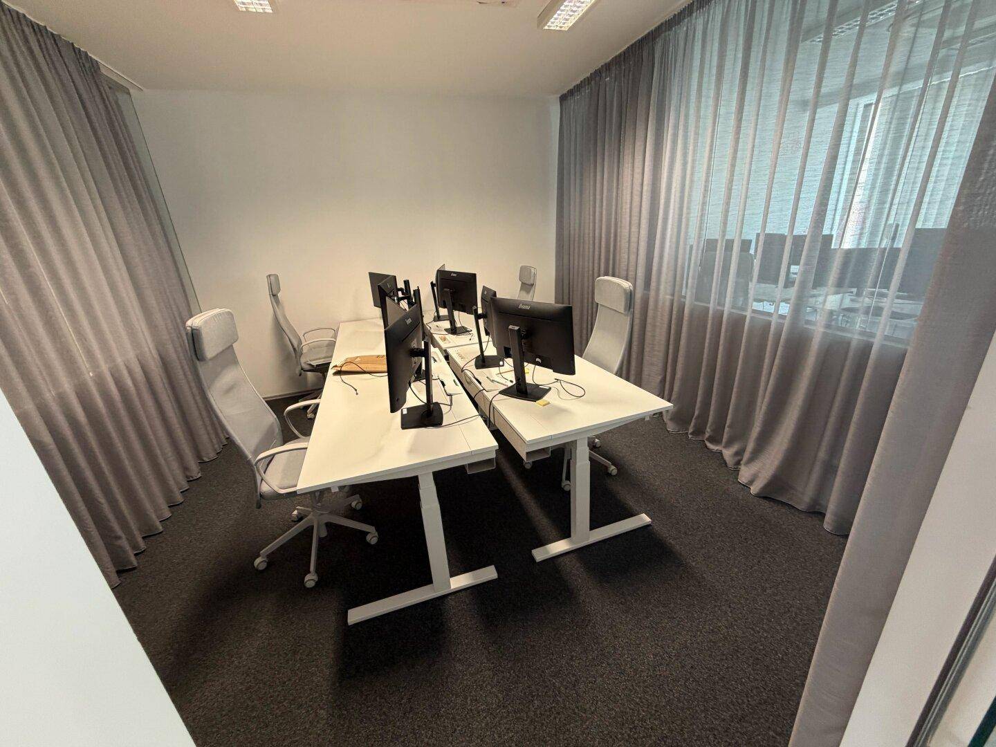 Modernes Büro mit mehreren Arbeitsplätzen, höhenverstellbaren Schreibtischen und grauen Vorhängen an den Fenstern.