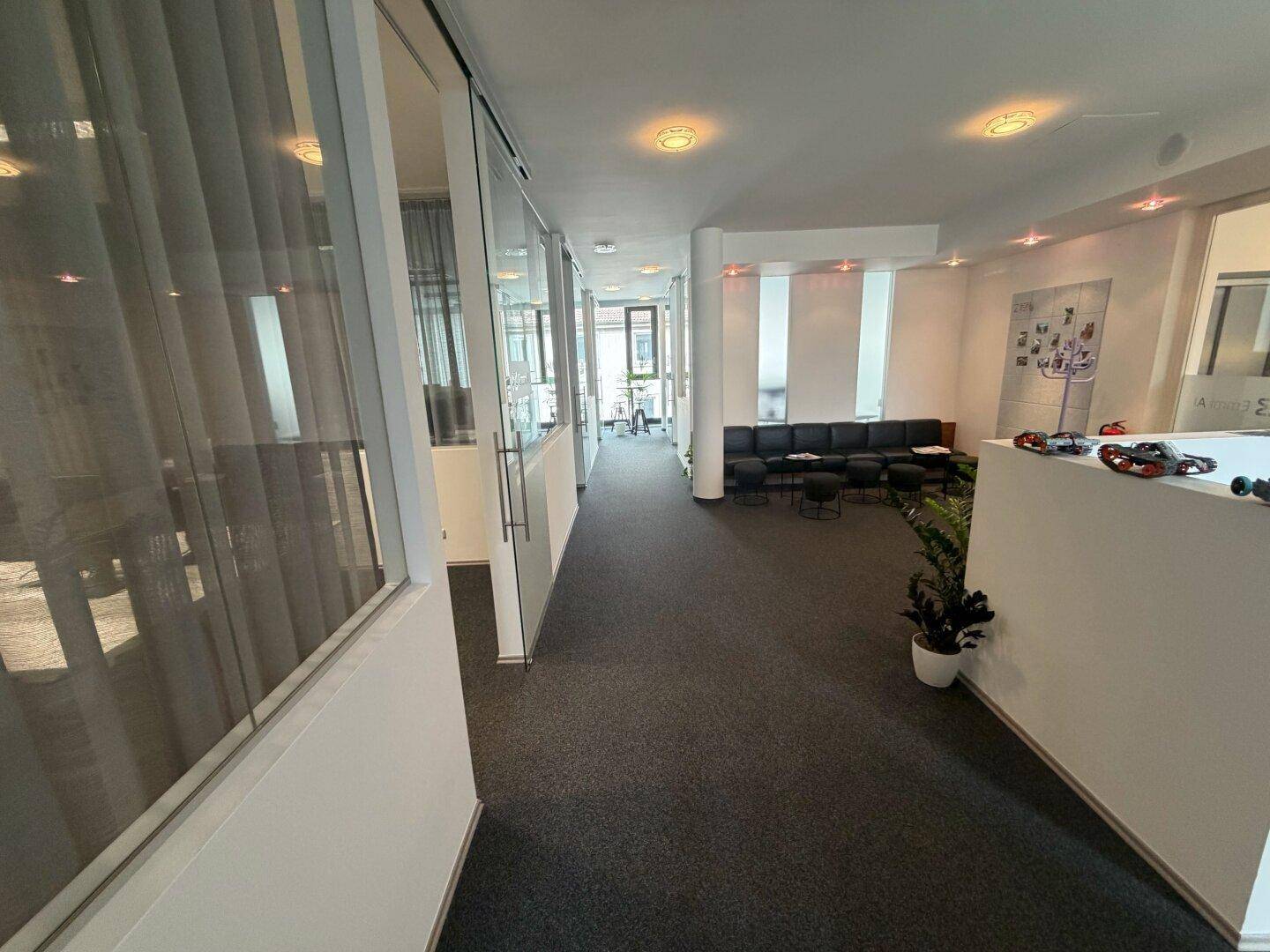 Heller Bürogang mit Teppichboden, Glaswänden und einem Wartebereich mit dunklen Sofas im Hintergrund.