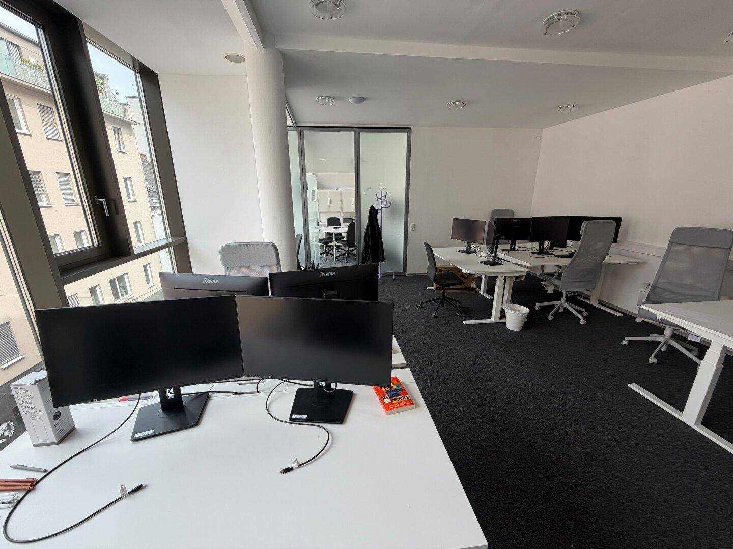 Geräumiges Büro mit mehreren Arbeitsplätzen, großen Fenstern und Blick auf die Umgebung und andere Gebäude.