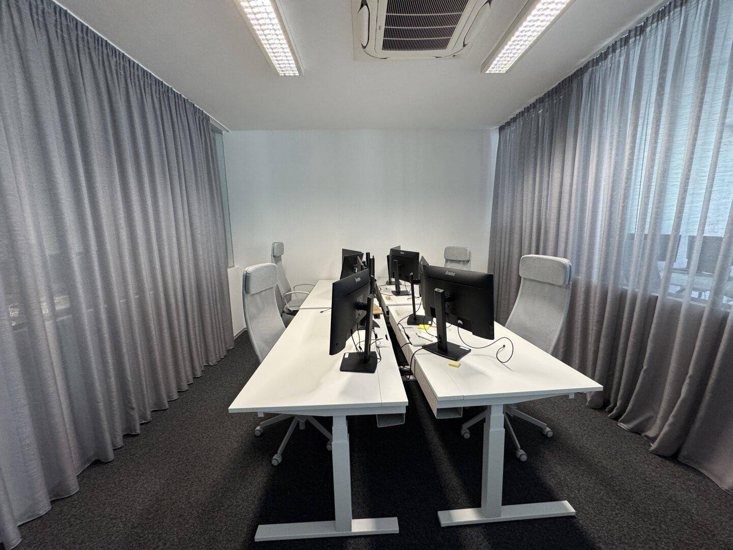 Modernes Büro mit mehreren Arbeitsplätzen, höhenverstellbaren Schreibtischen und grauen Vorhängen an den Fenstern.