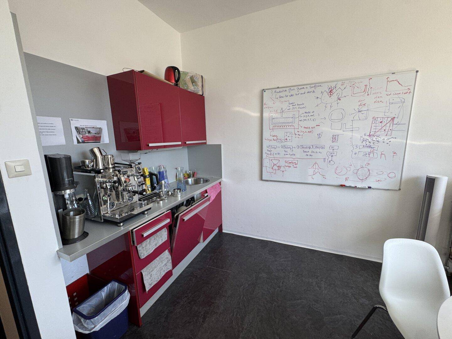 Kleine Küchenzeile mit roten Schränken, Spüle und Kaffeemaschine in einem Bürobereich für Mitarbeiter.
