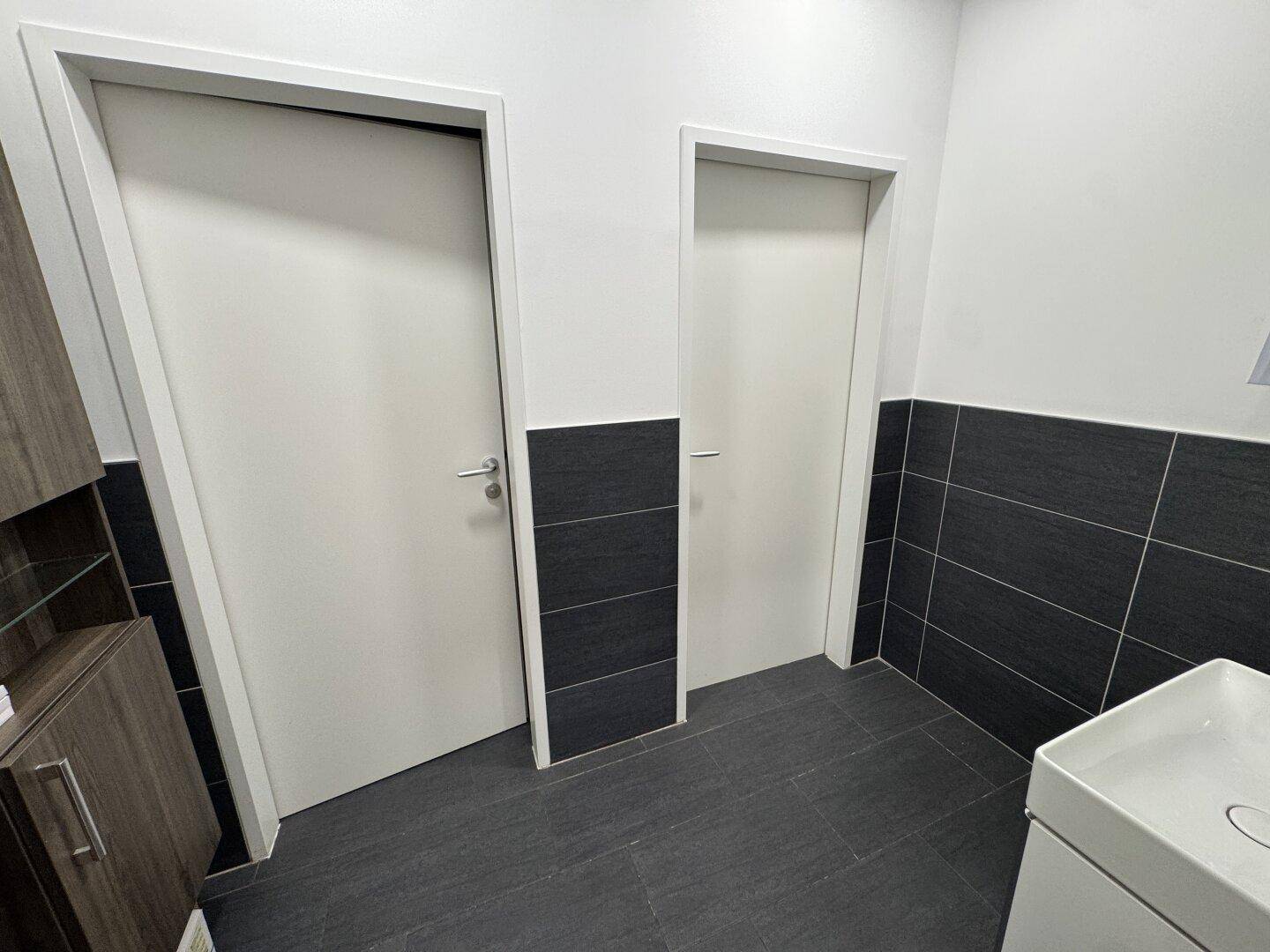 Toilettenbereich mit dunklen Fliesen und zwei weißen Türen, die zu den Kabinen führen.
