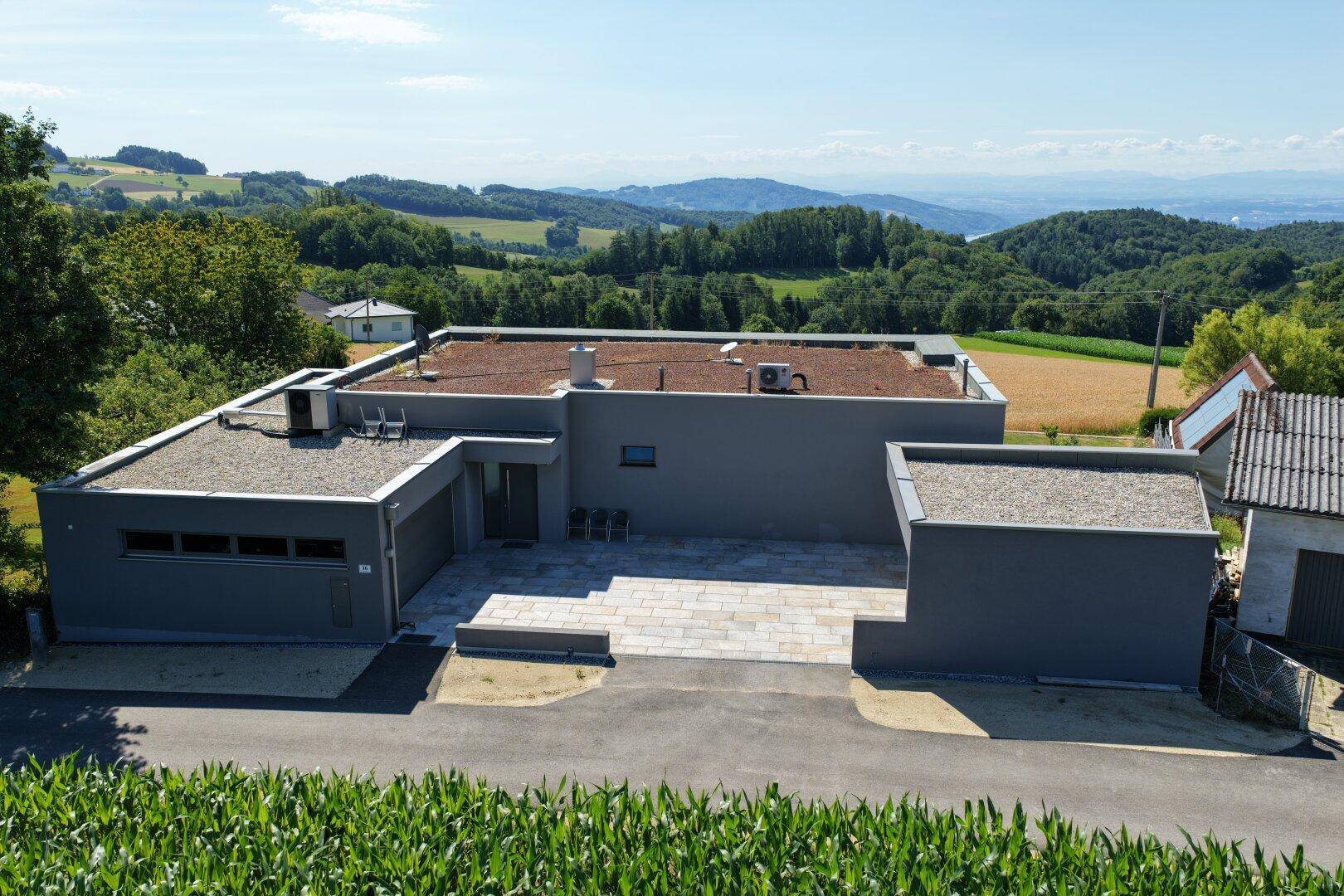 Rückansicht eines modernen Hauses mit Flachdach und gepflastertem Vorplatz, umgeben von ländlicher Idylle.