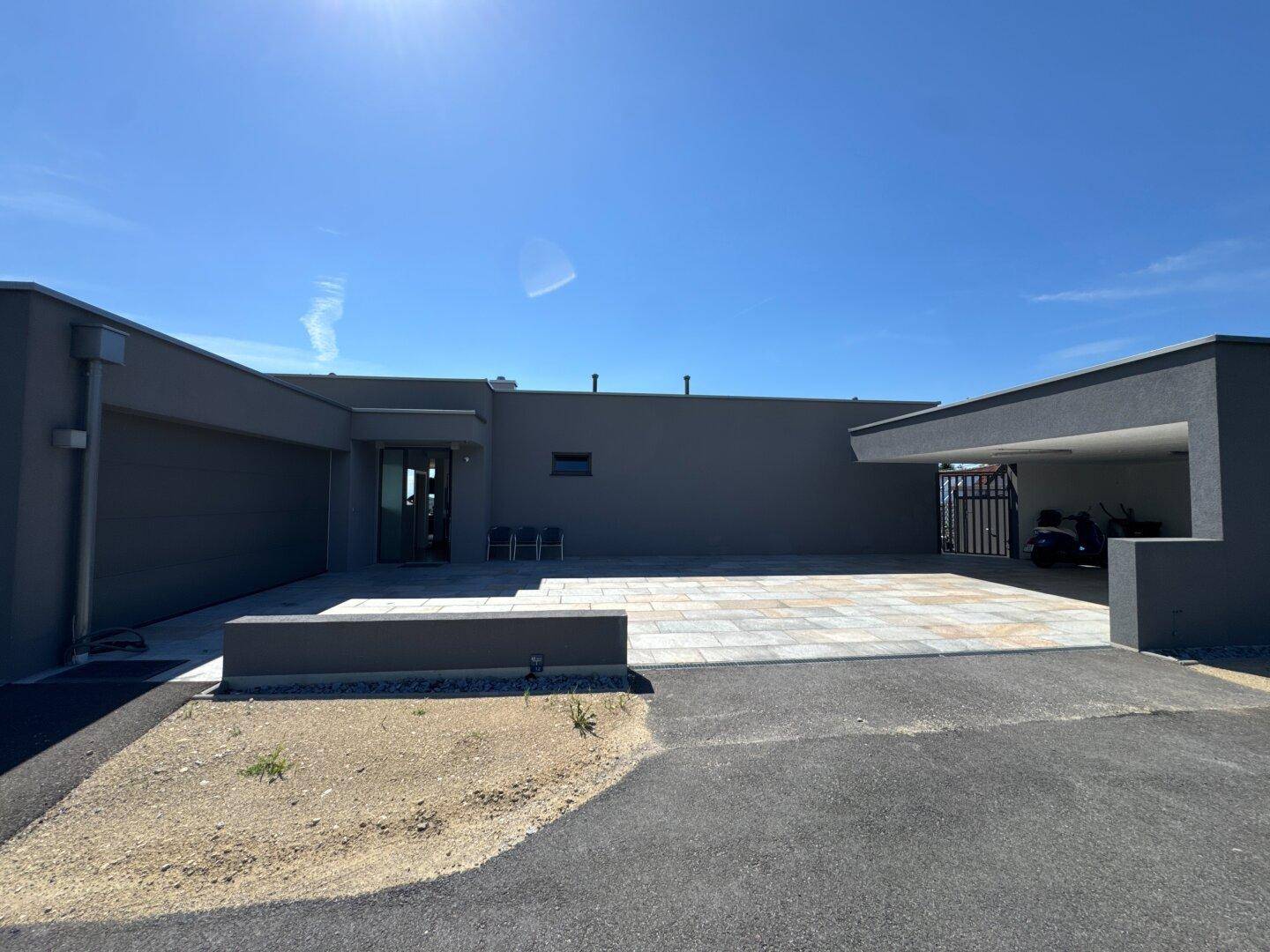 Moderner Eingangsbereich eines Hauses mit gepflastertem Vorplatz und Carport unter strahlend blauem Himmel.