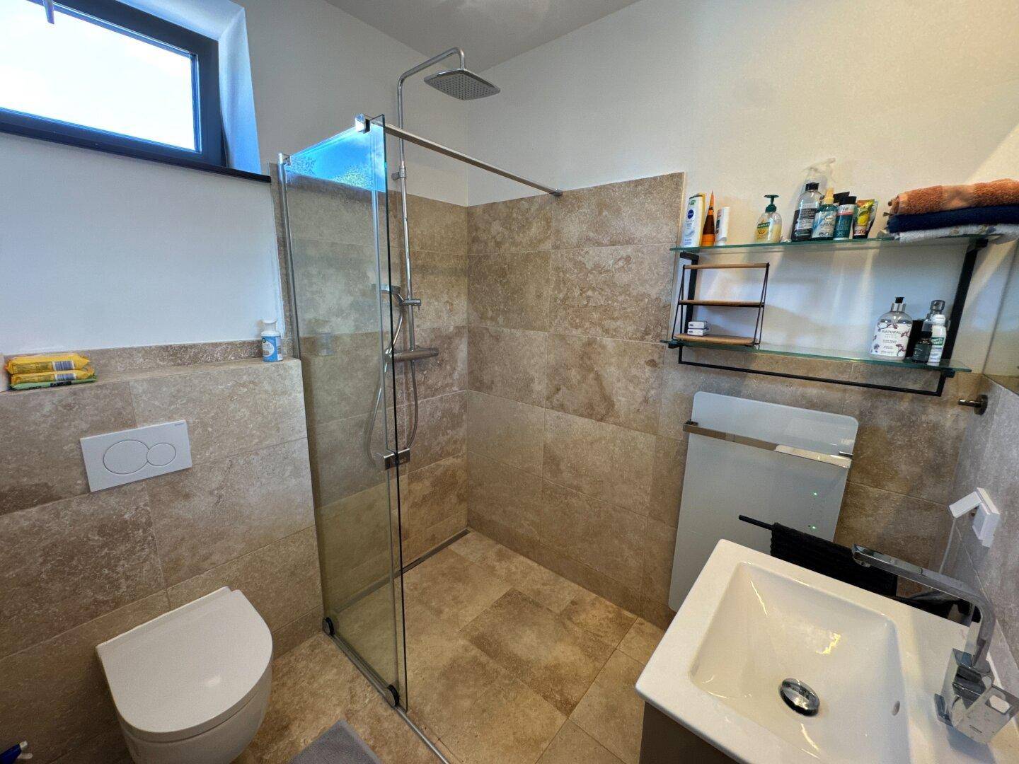 Modernes Badezimmer mit begehbarer Dusche, Wand-WC und Waschbecken, gefliest in Natursteinoptik.