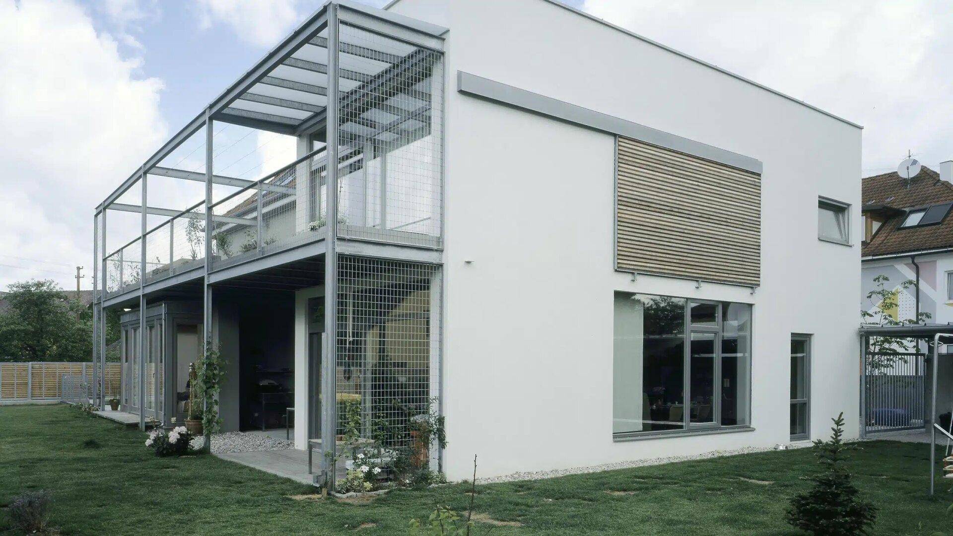 Moderne Hausfassade mit großen Fenstern, Holzlamellen und Gartenbereich.