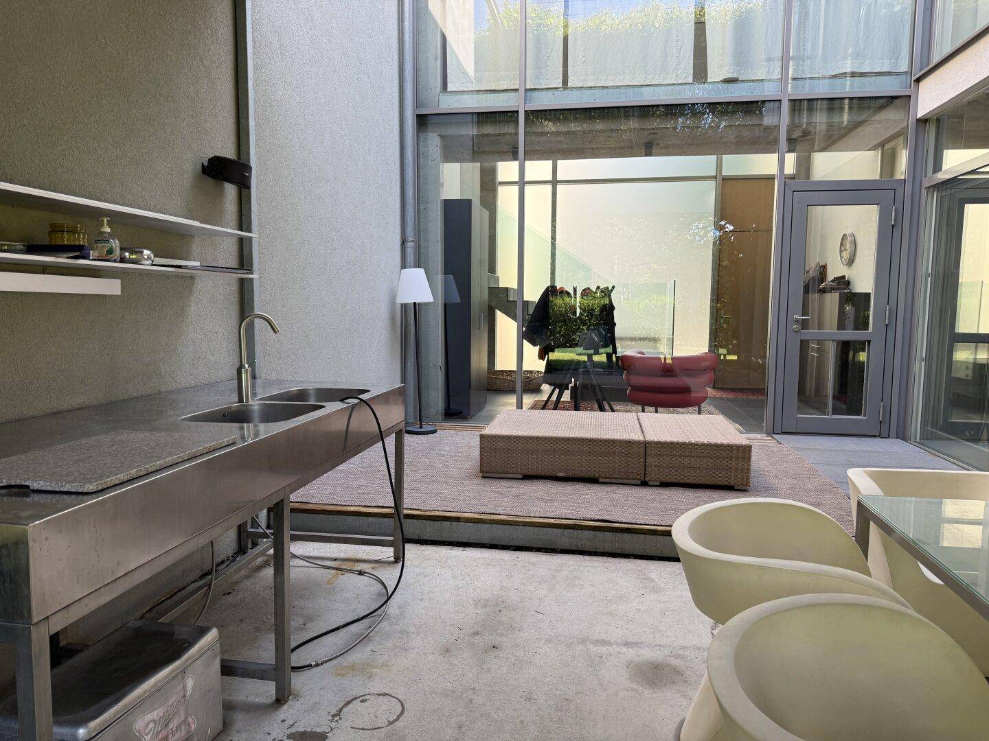 Überdachte Terrasse mit Außenküche und Sitzbereich, verbunden mit dem Innenraum durch Glasfronten.