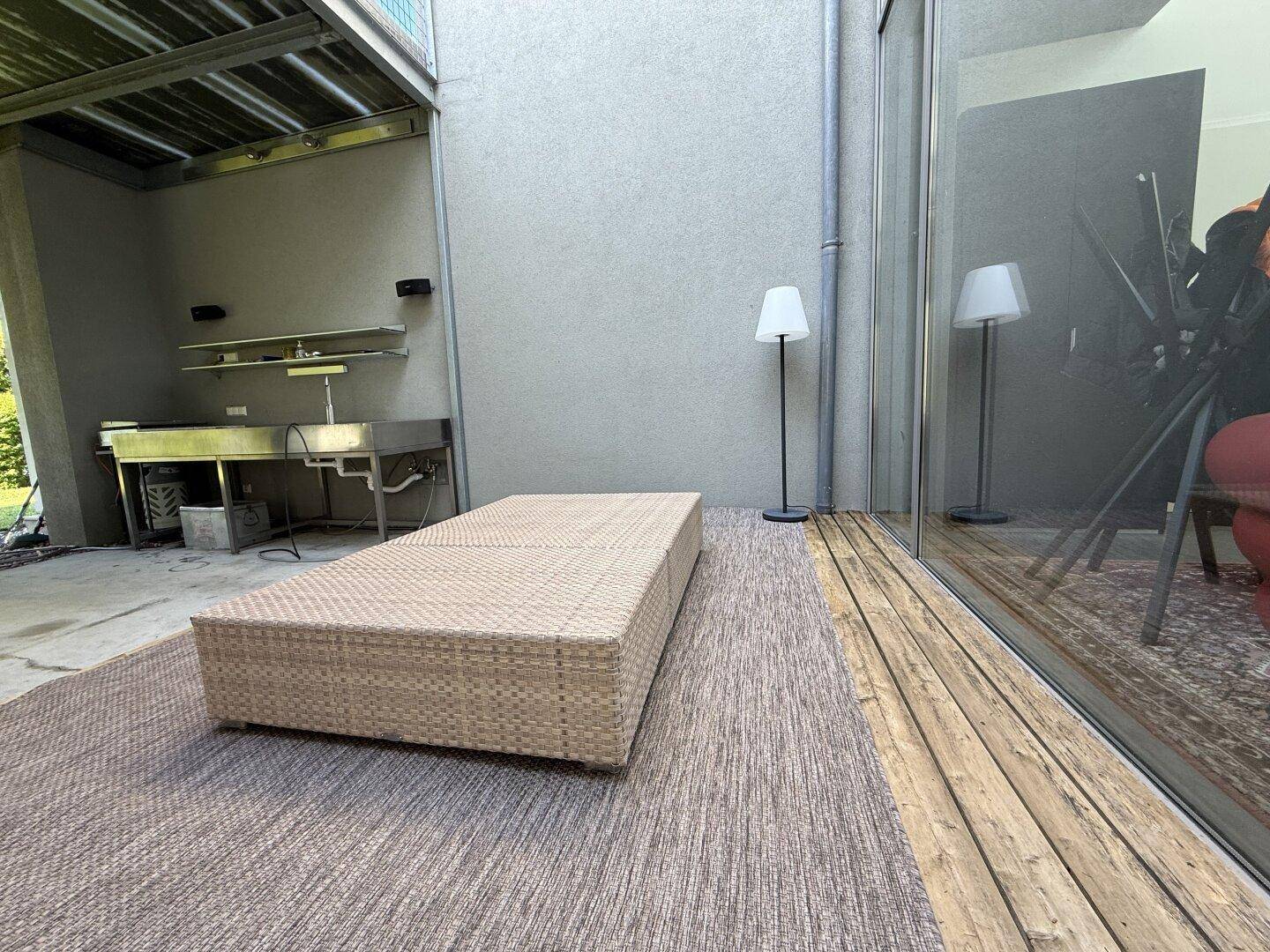 Entspannungsbereich auf der Terrasse mit Rattanliege und Holzboden, angrenzend an eine Glasfront.