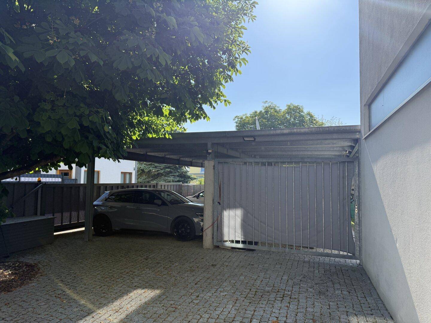 Überdachter Carport mit gepflasterter Einfahrt und schattenspendenden Bäumen.