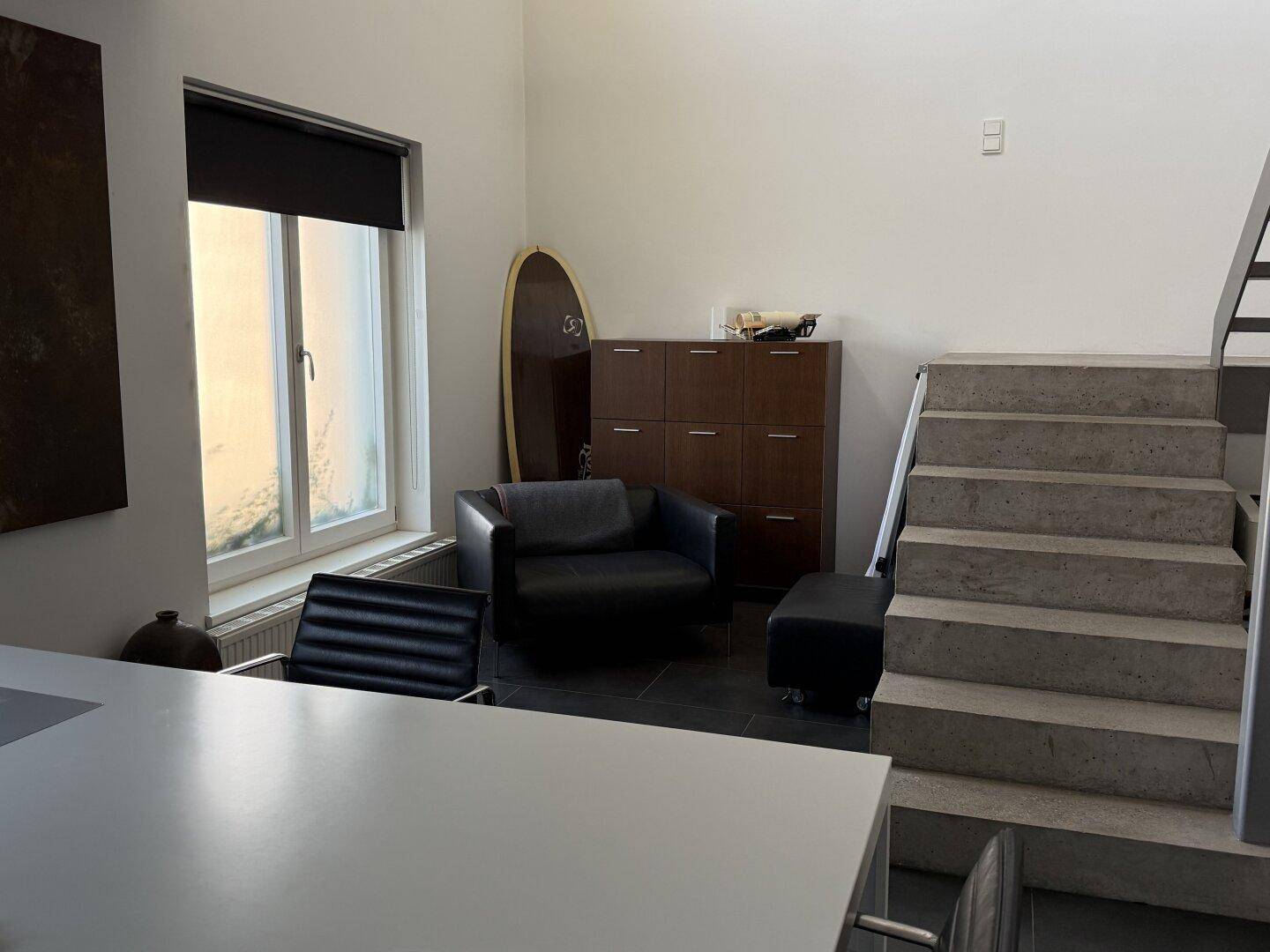 Wohnbereich mit dunklem Fliesenboden, moderner Treppe und großem Fenster für viel Licht.