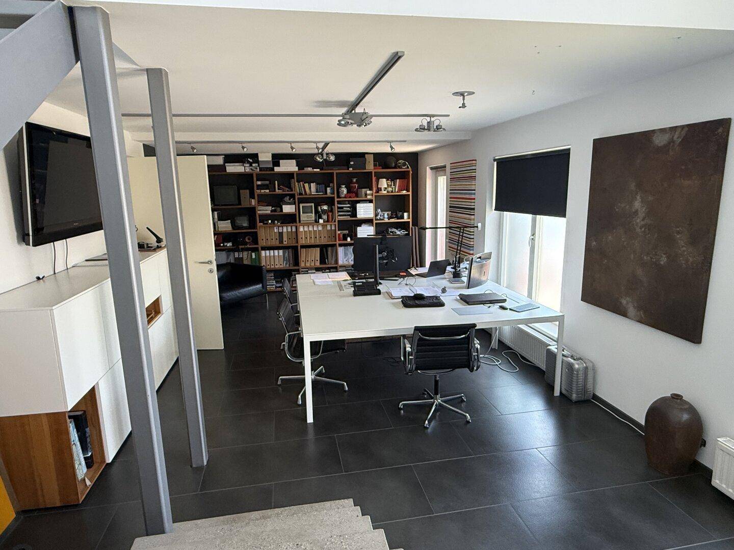 Modernes Büro mit großem Schreibtisch, Fernseher und offener Treppe, die den Raum strukturiert.